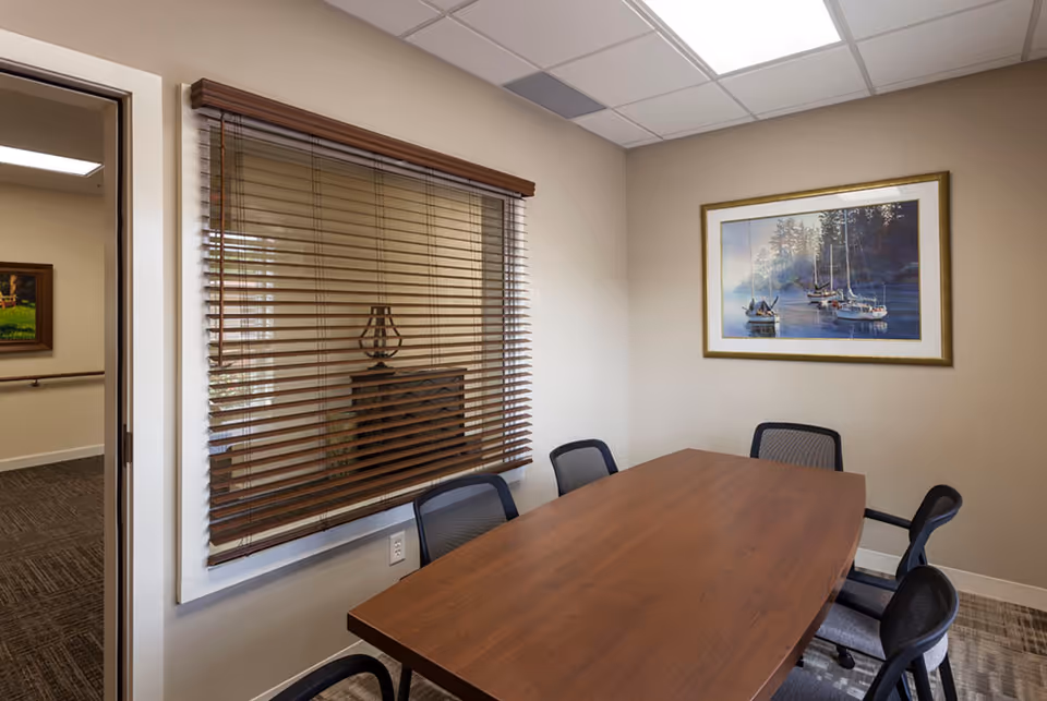 A conference-style room with a wooden table, chairs, blinds, and a framed landscape artwork.