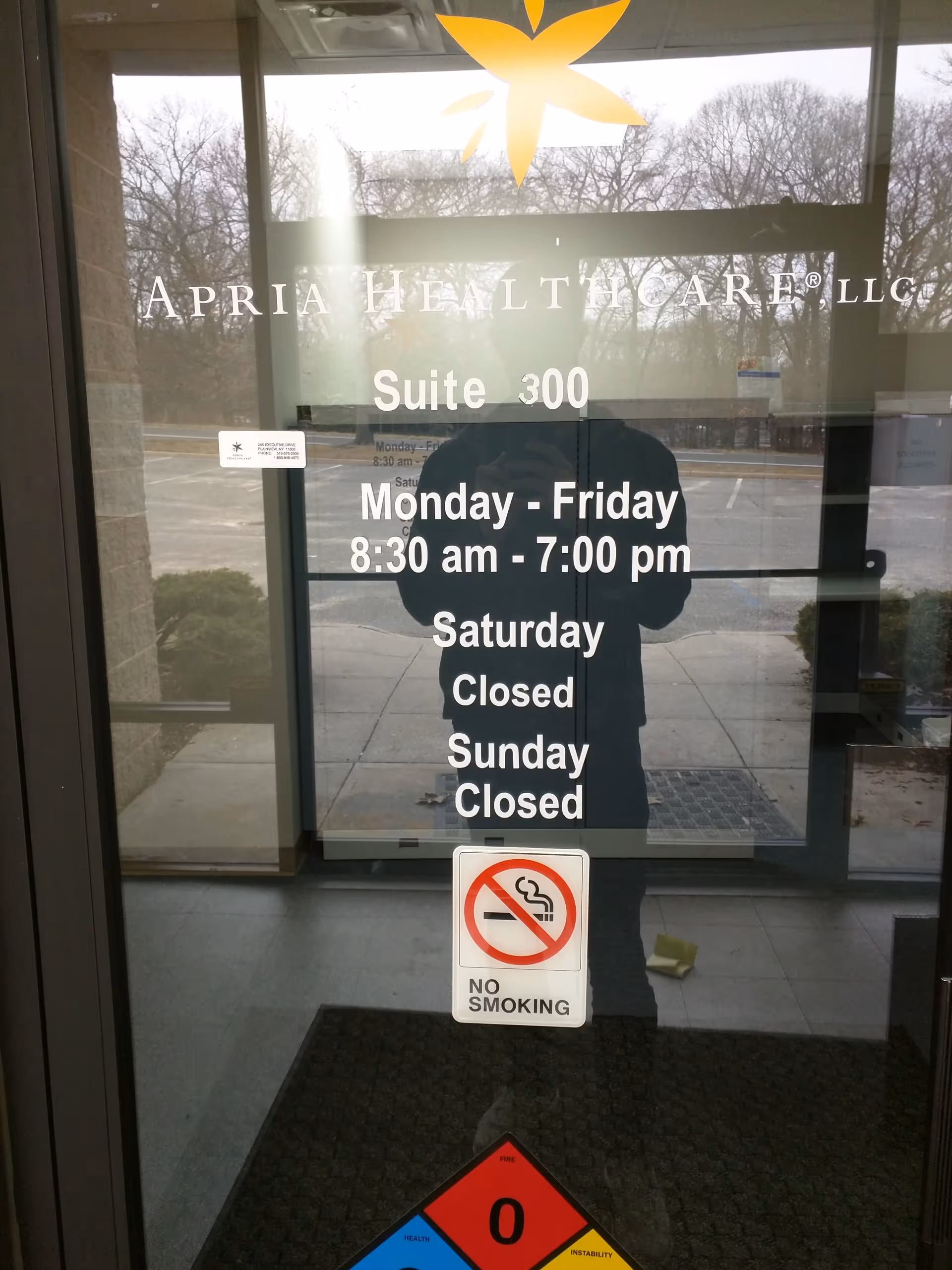 Glass entrance door displays Apria Healthcare Suite 300 hours and no-smoking sign.