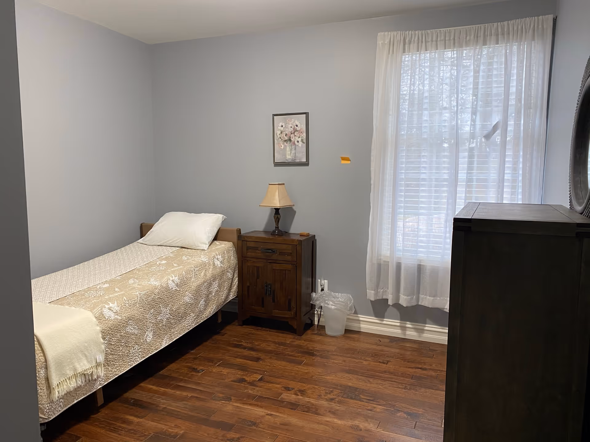 Small bedroom in a senior living facility with a single bed, dresser, lamp, and curtains.