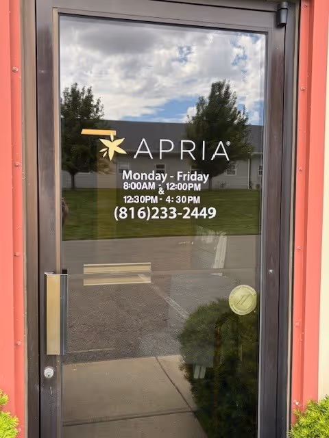 Glass door with APRIA logo and hours, reflecting trees and sidewalk outside.
