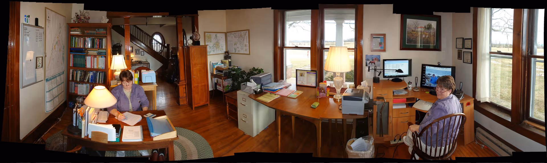 Panoramic view of a cozy office with desks, computers, bookshelves, and two workers.