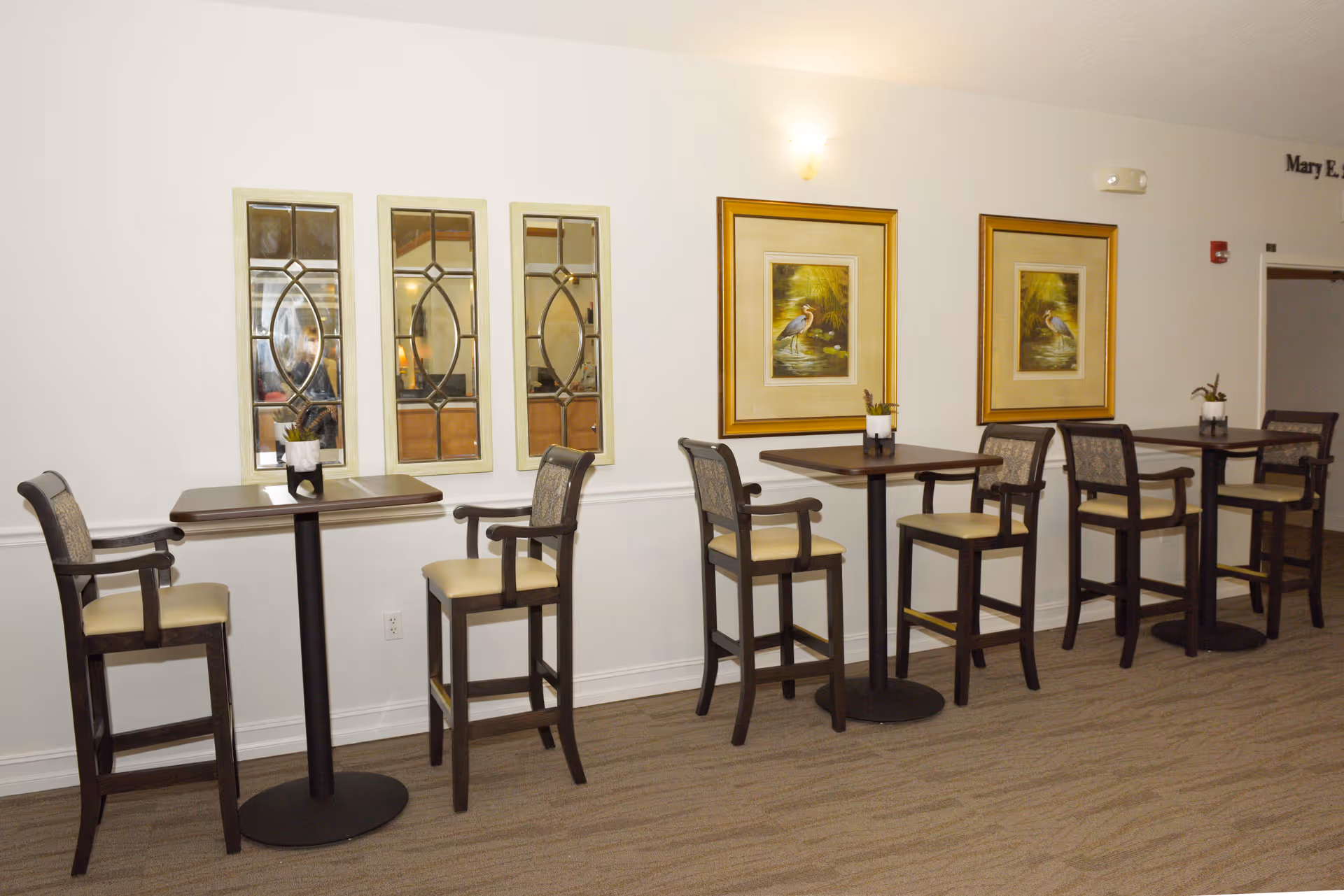 Interior hospice lounge with high-top tables, chairs, and framed wall art along a white wall.