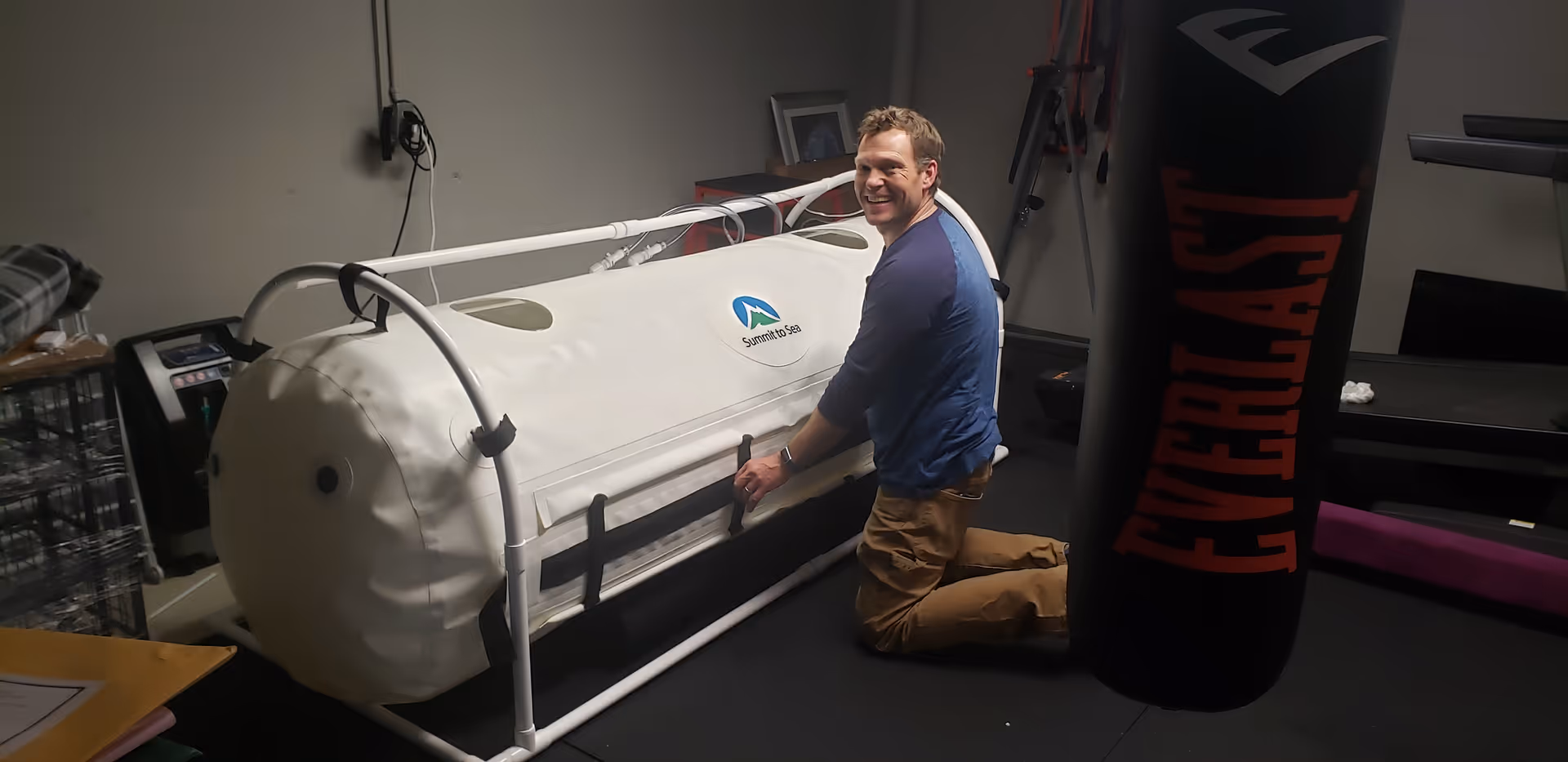 A man kneels beside a white hyperbaric chamber in a small, equipment-filled therapy room.