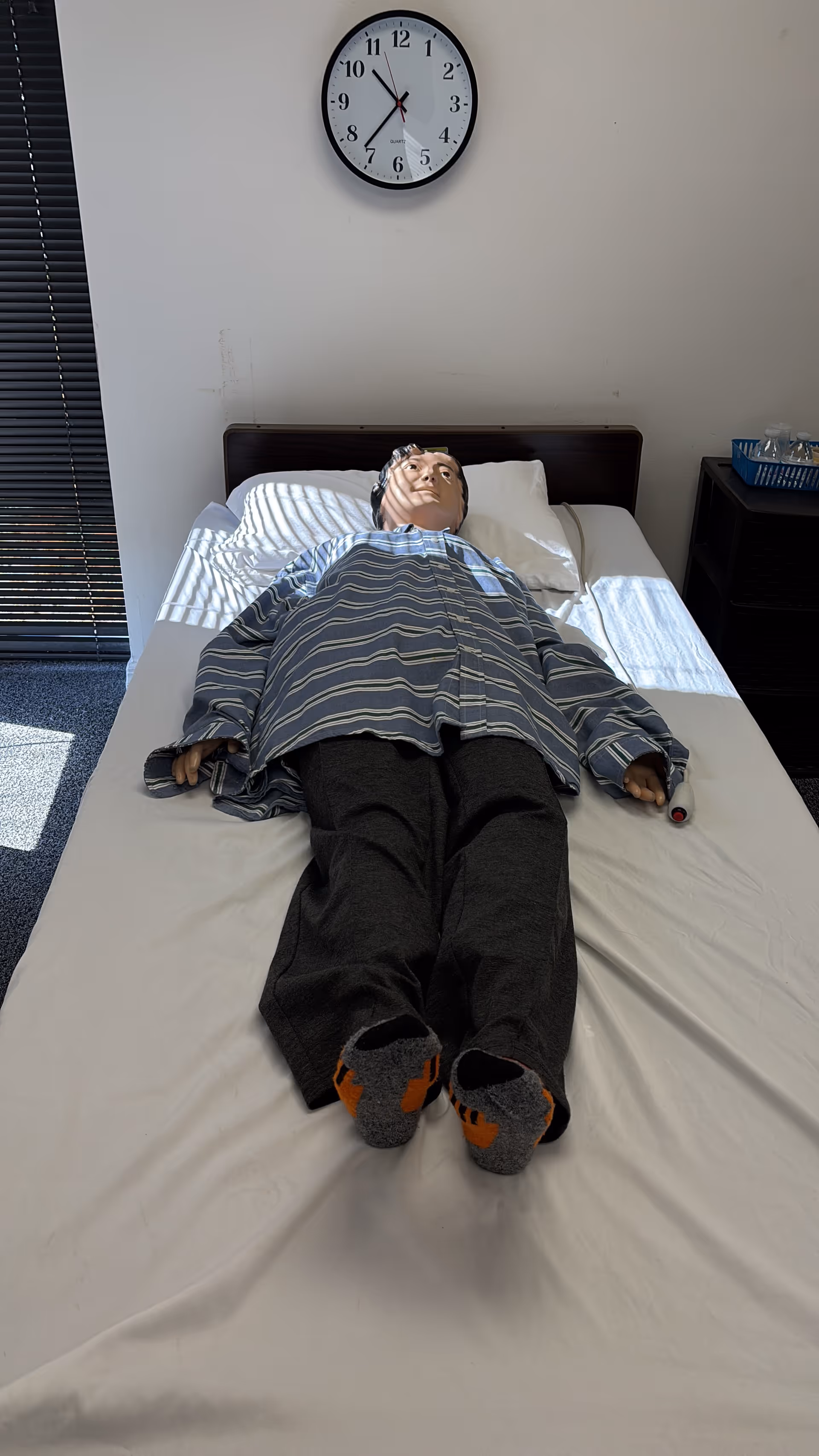 A medical training mannequin lies on a bed inside a clinical training room.