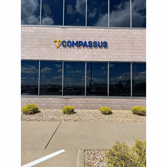 Front exterior of Compassus facility with logo, large windows, and trimmed shrubs.