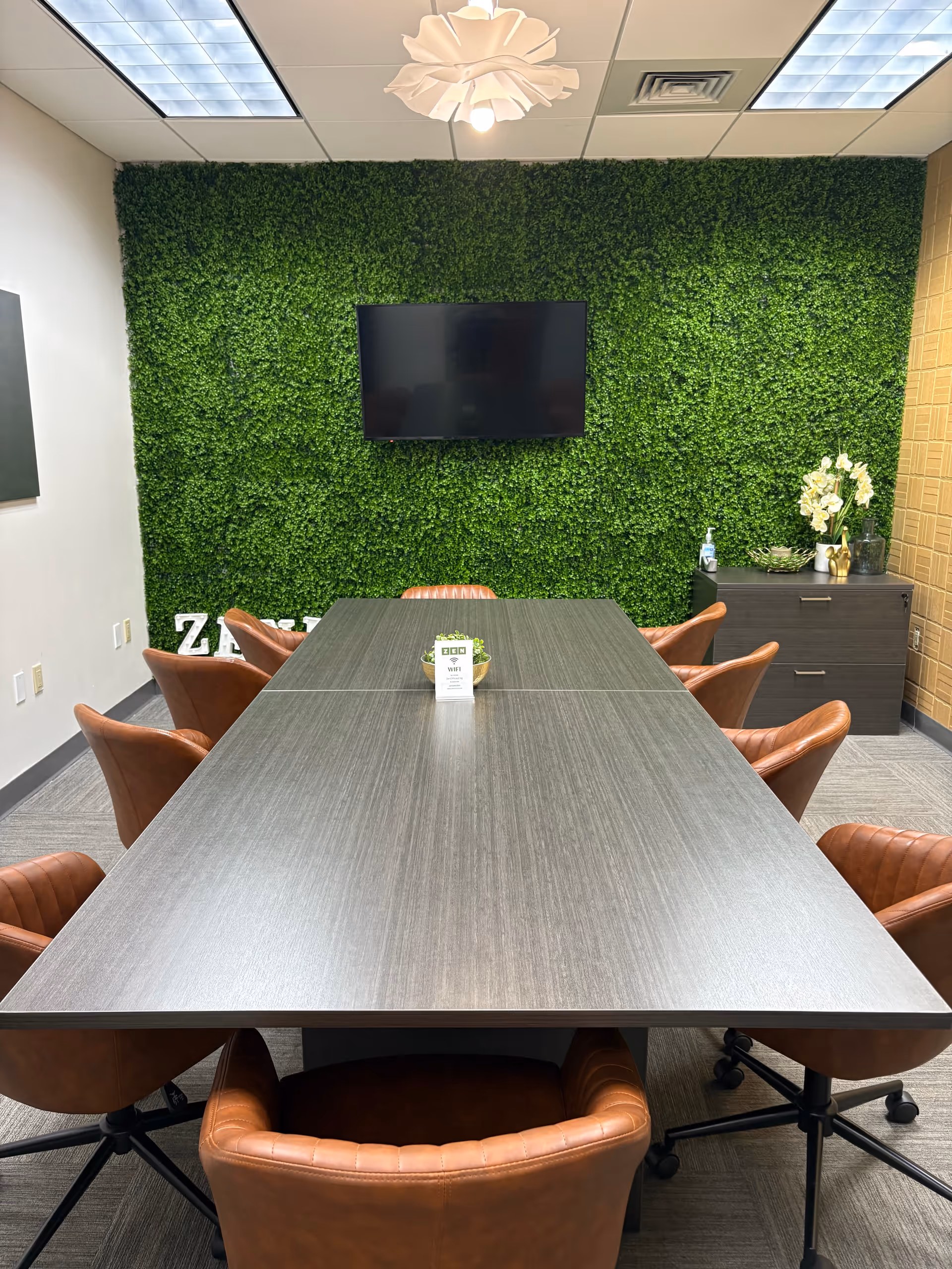 A modern conference room with a long table, brown chairs, and a green plant wall.