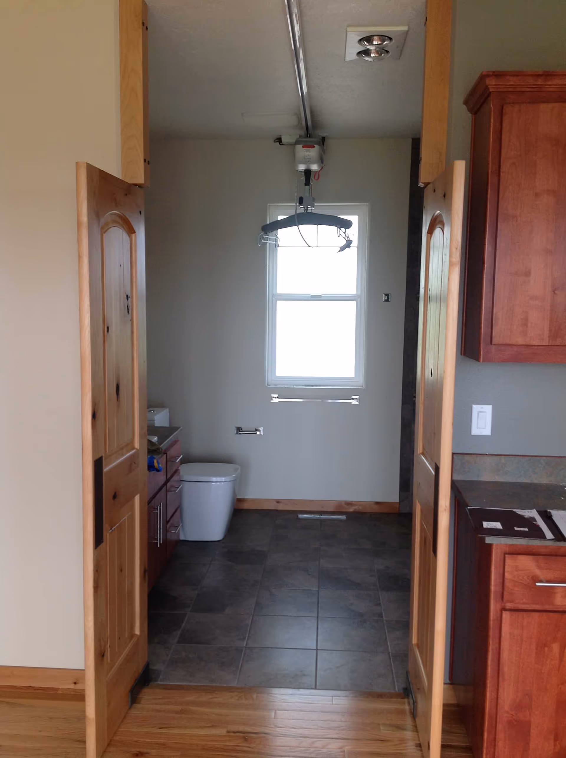 Narrow bathroom through doorway with toilet, cabinets, window, and ceiling hoist.