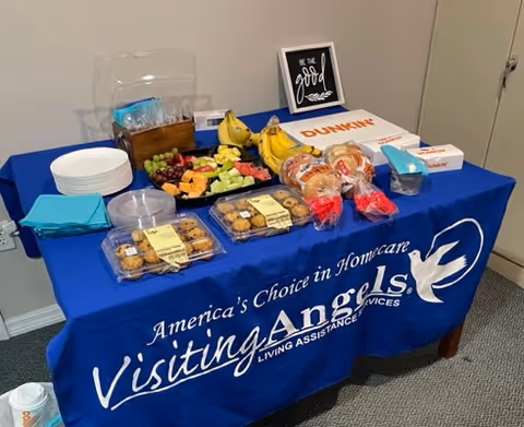 Blue Visiting Angels table with snacks, fruit, pastries, and plates in a hallway.