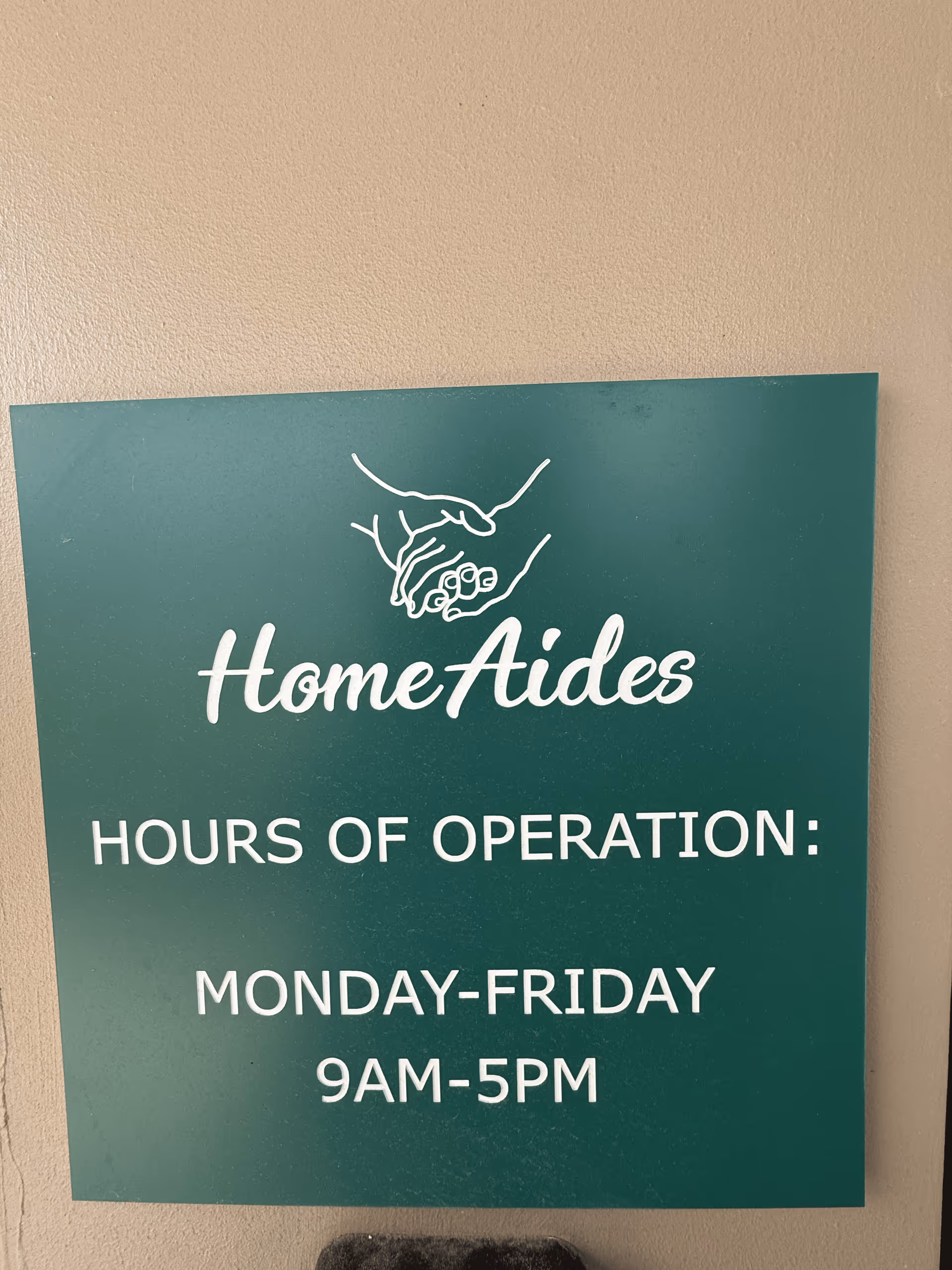 Green HomeAides sign detailing hours of operation mounted on a beige wall.