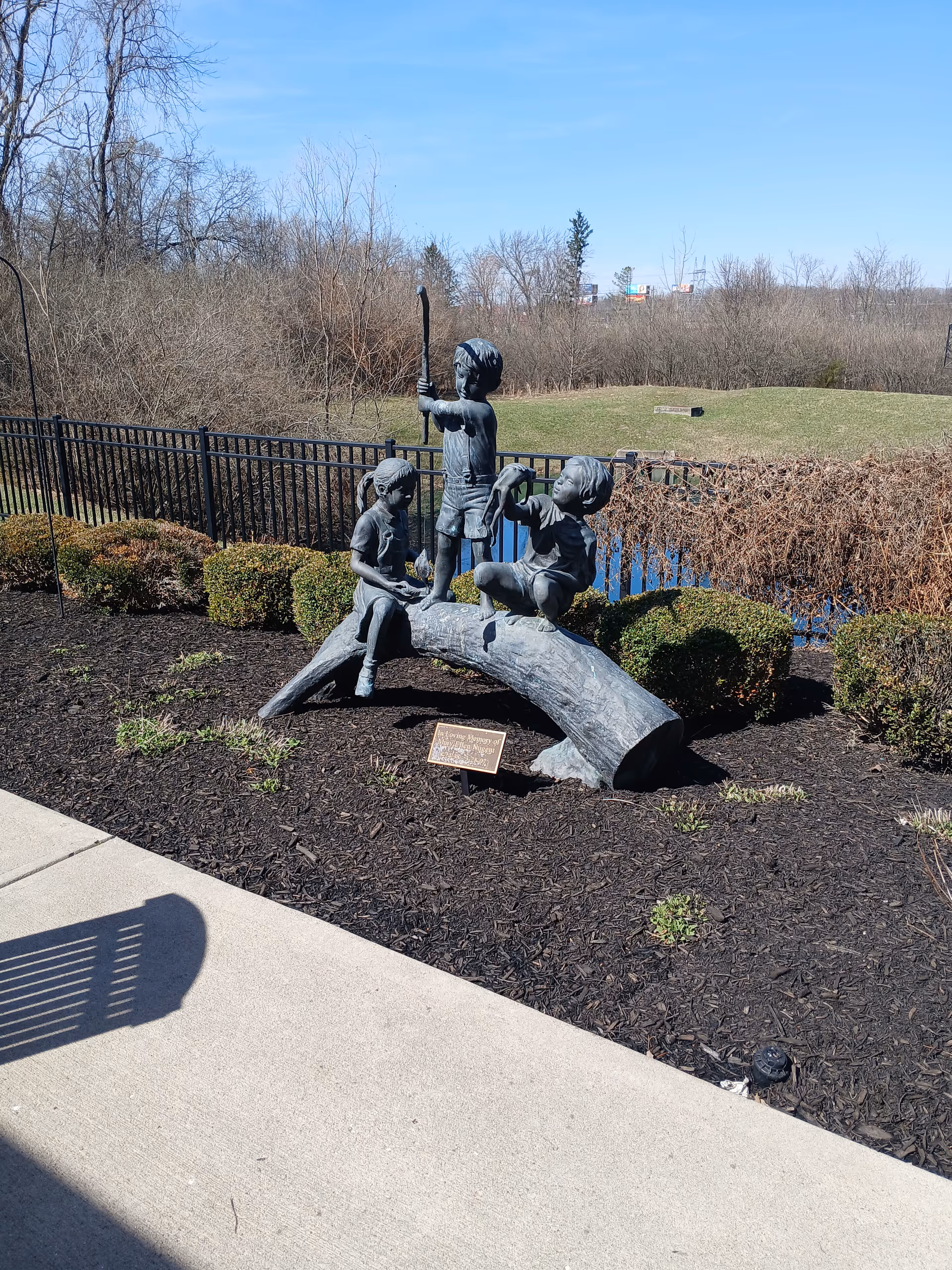 Outdoor sculpture of three children on a log in a landscaped area near a fence
