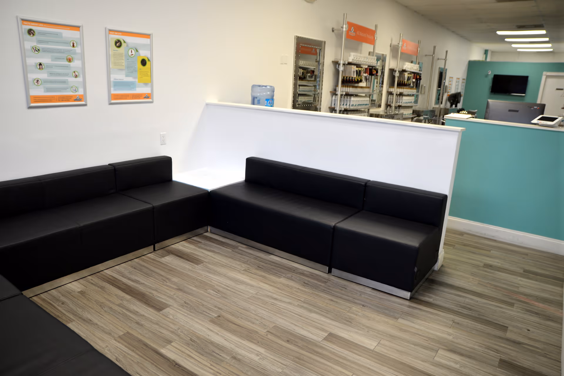 Waiting area with black L-shaped couches, white walls, posters, and a teal reception desk.
