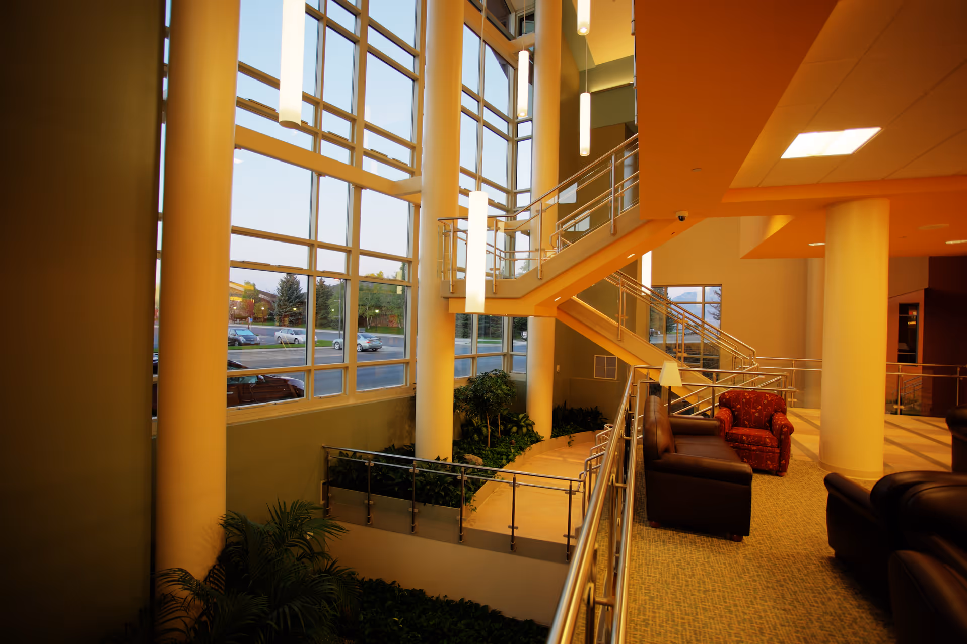 Bright hospital atrium with large windows, stairs, railings, seating, and plants