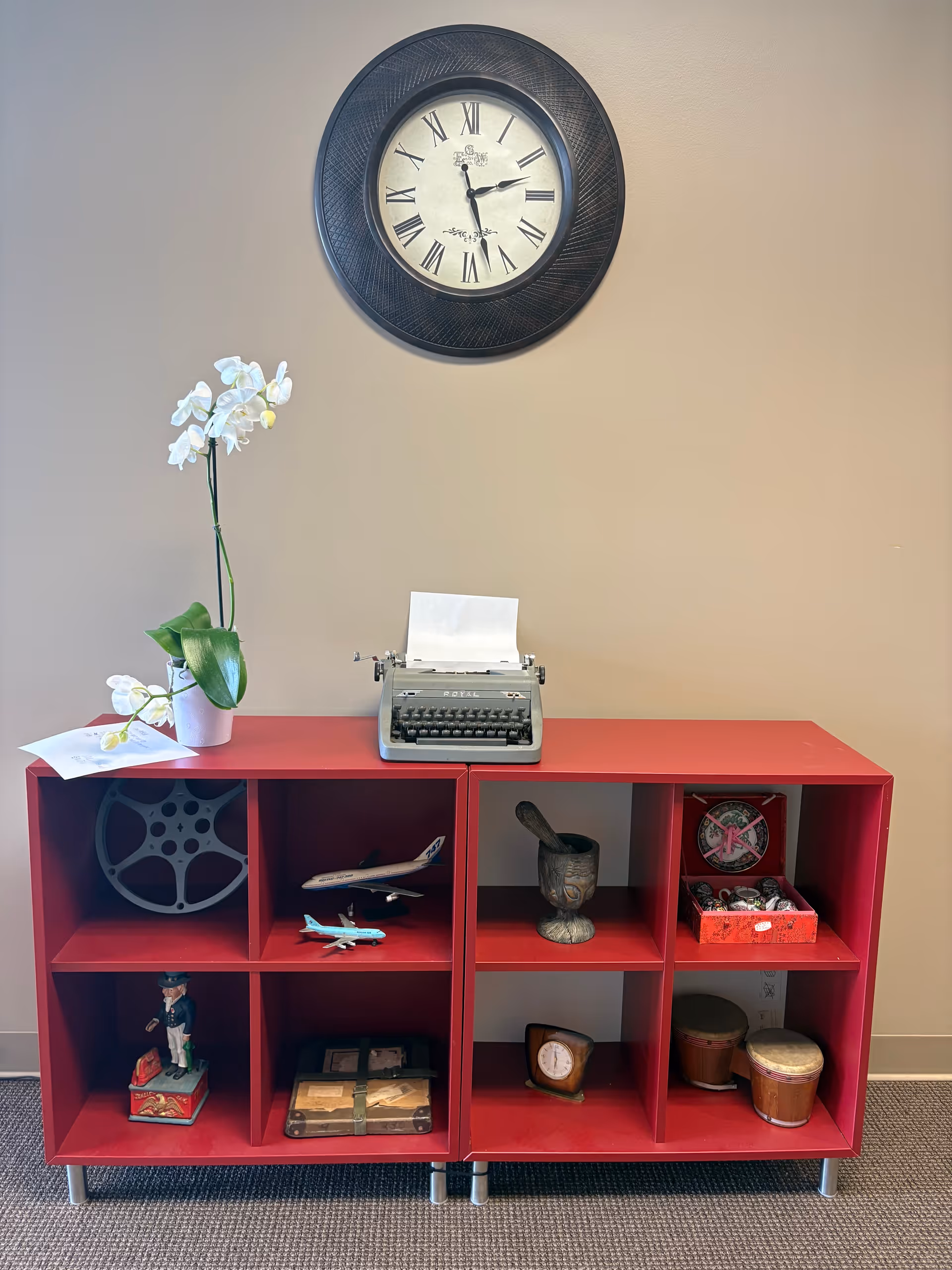 Red cubby shelf with vintage typewriter, orchid, and decorative items beneath a large wall clock.