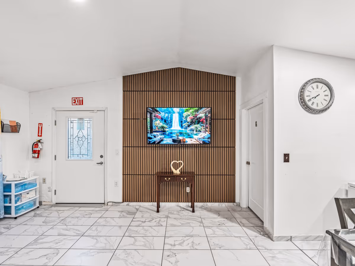 Lobby-like interior with wood-paneled wall, wall-mounted TV, console table, exit door, and marble floor.
