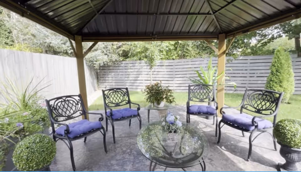 Outdoor gazebo seating with metal chairs and a glass table under a shaded pavilion.