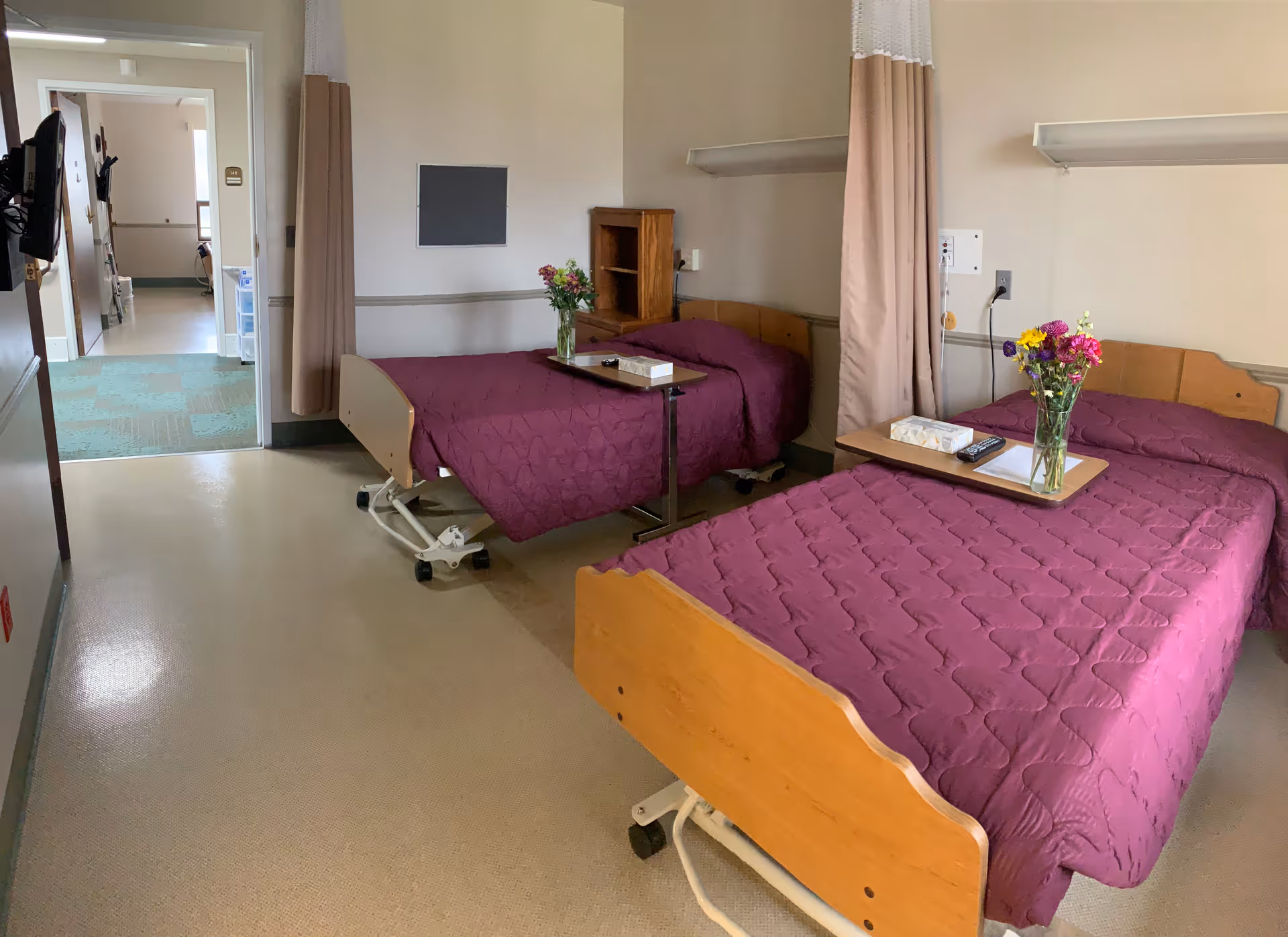 Two hospital beds with purple quilts, side tables, and flowers in a shared patient room.