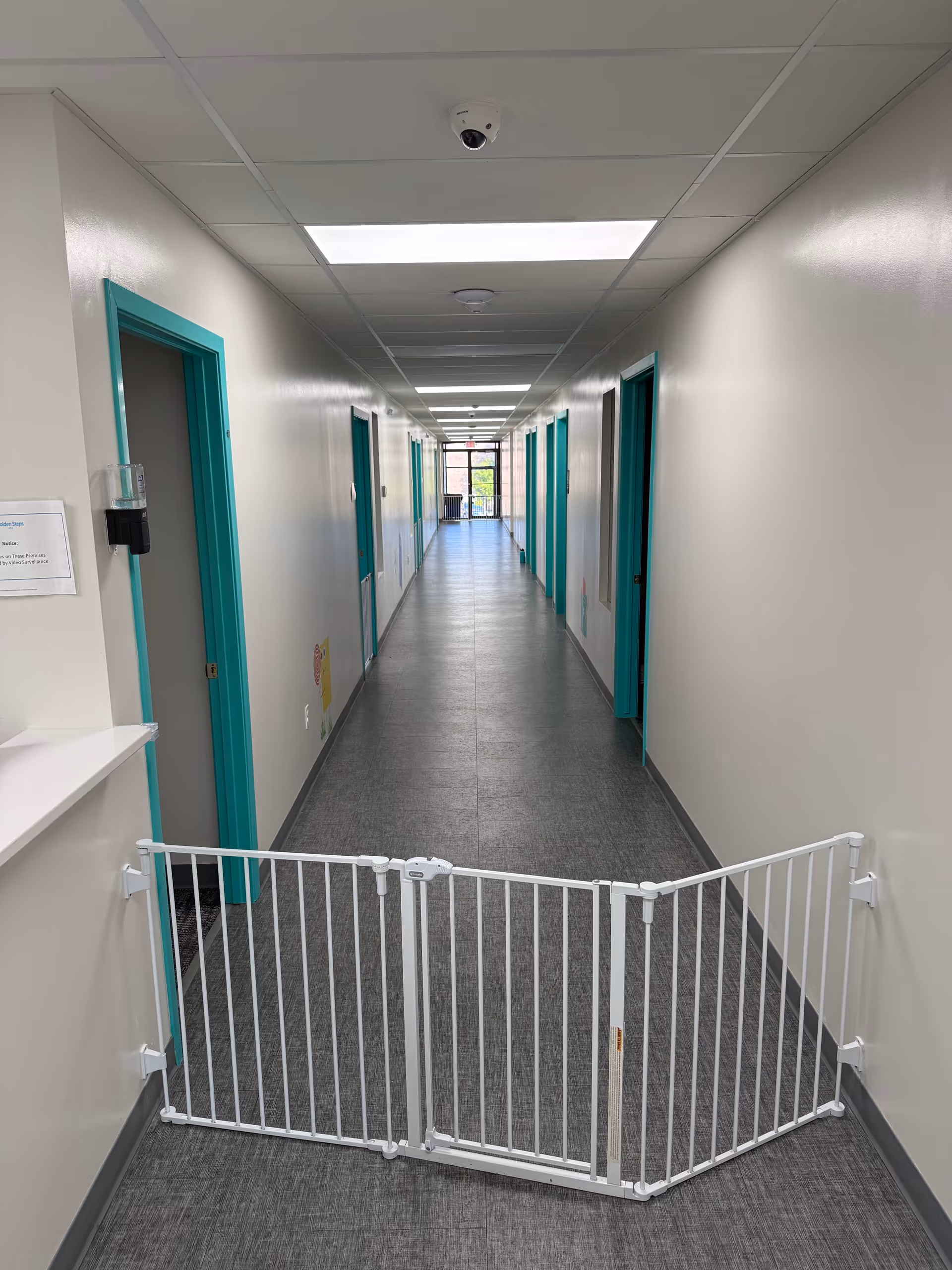 Interior hallway with teal door frames, ceiling lights, and a white safety gate across the corridor.