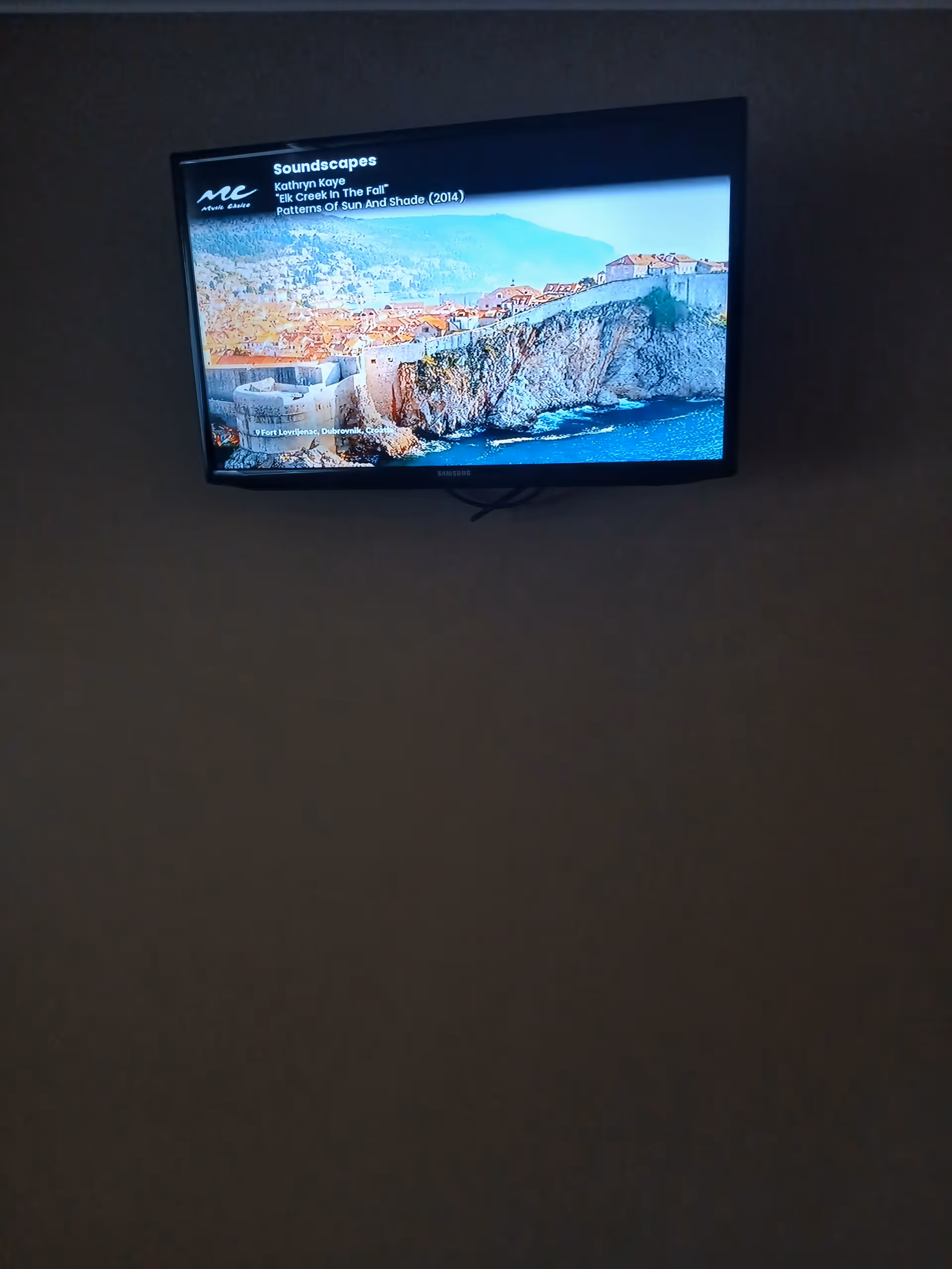Wall-mounted flat-screen TV displaying a coastal landscape inside a dim room.