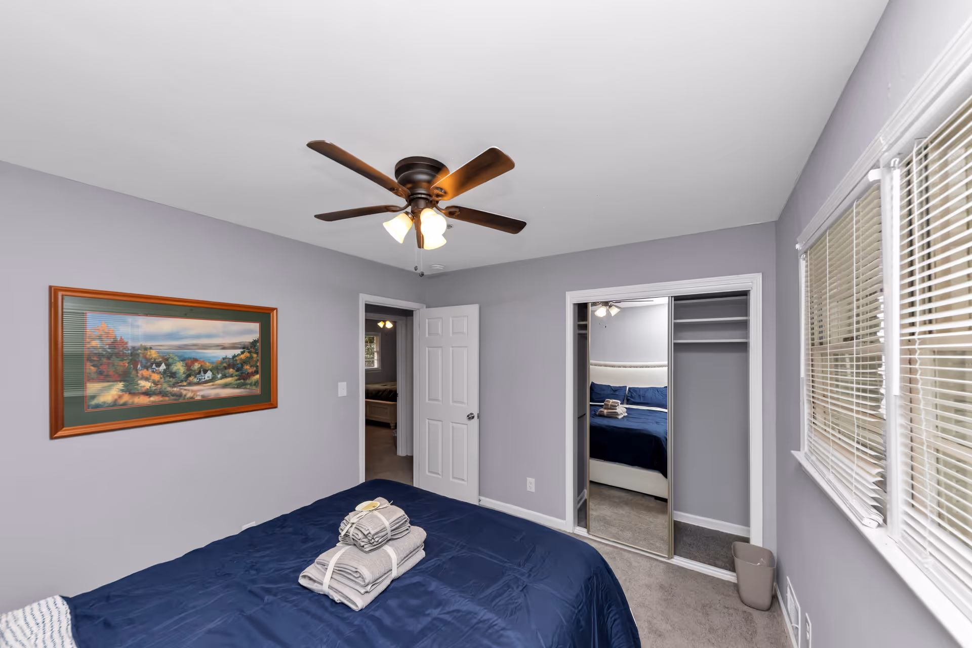 Purple-walled bedroom with a blue bedspread, ceiling fan, window blinds, and a mirrored closet.