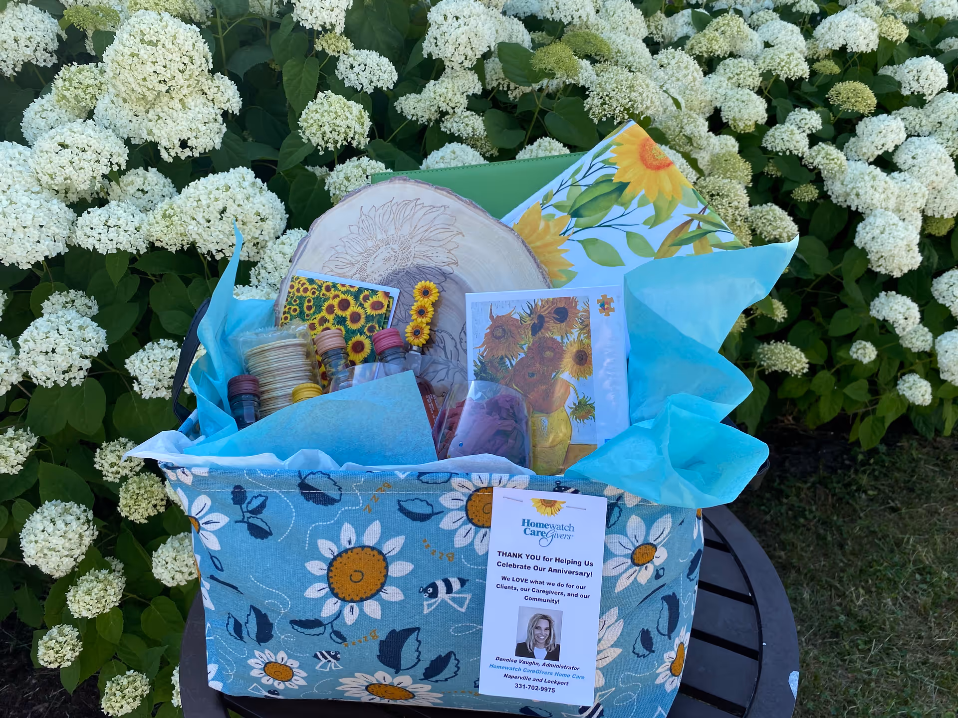 Gift basket on blue floral cloth sits on a table outside near white hydrangea bushes