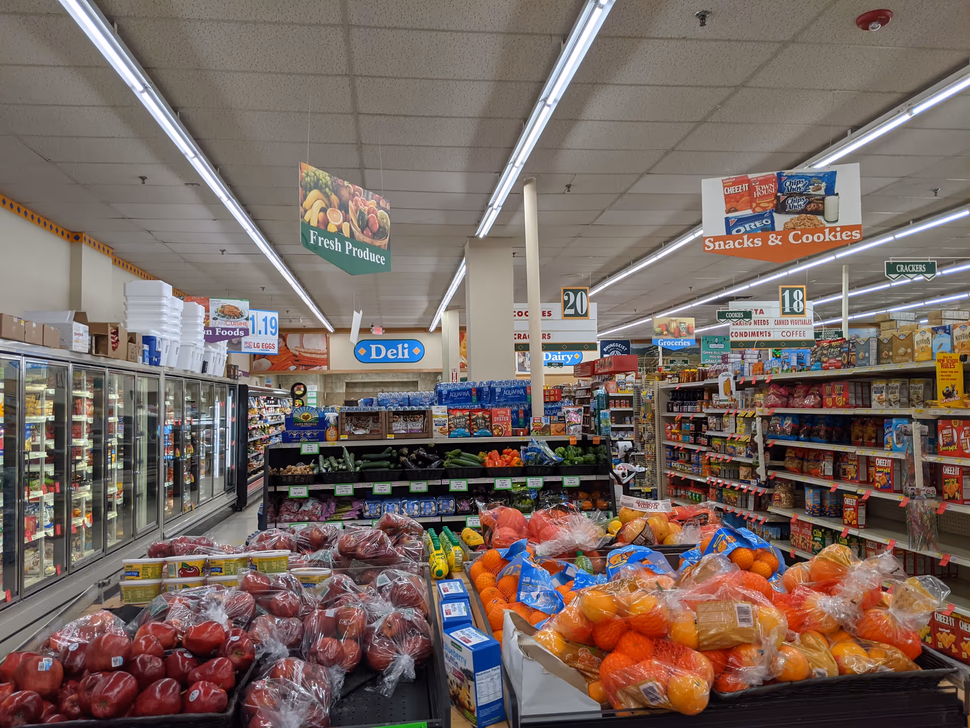 Wide grocery store aisle inside a facility, with produce and snack signs overhead.