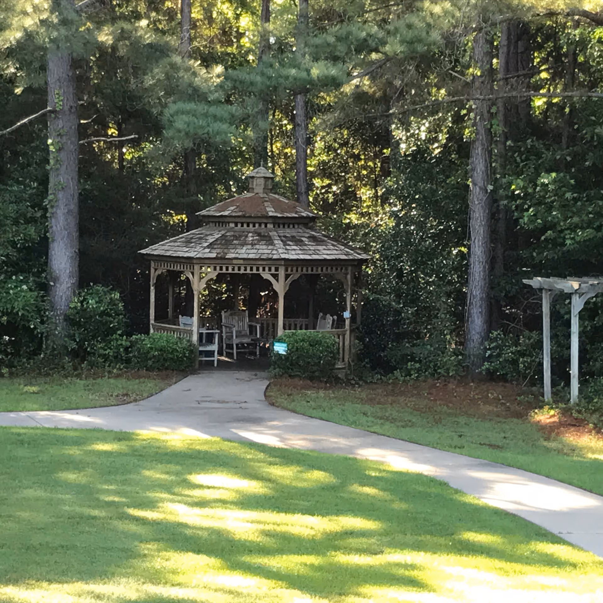 A wooden gazebo with chairs sits along a paved path in a shaded garden.