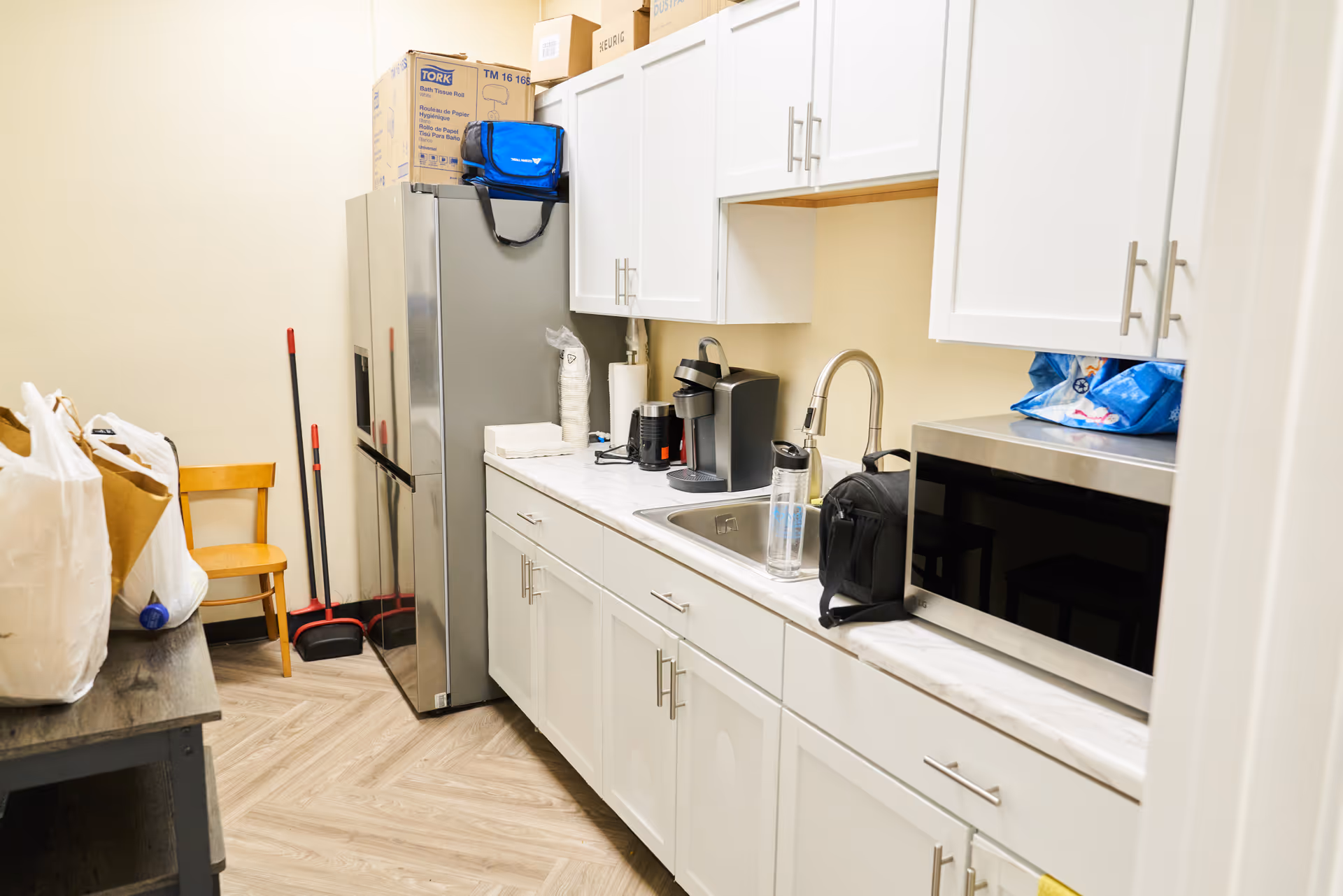 A compact white kitchen with refrigerator, cabinets, sink, microwave, and cleaning supplies.