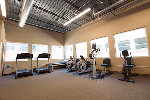 Interior fitness room with treadmills, stationary bikes, ellipticals, and large windows.