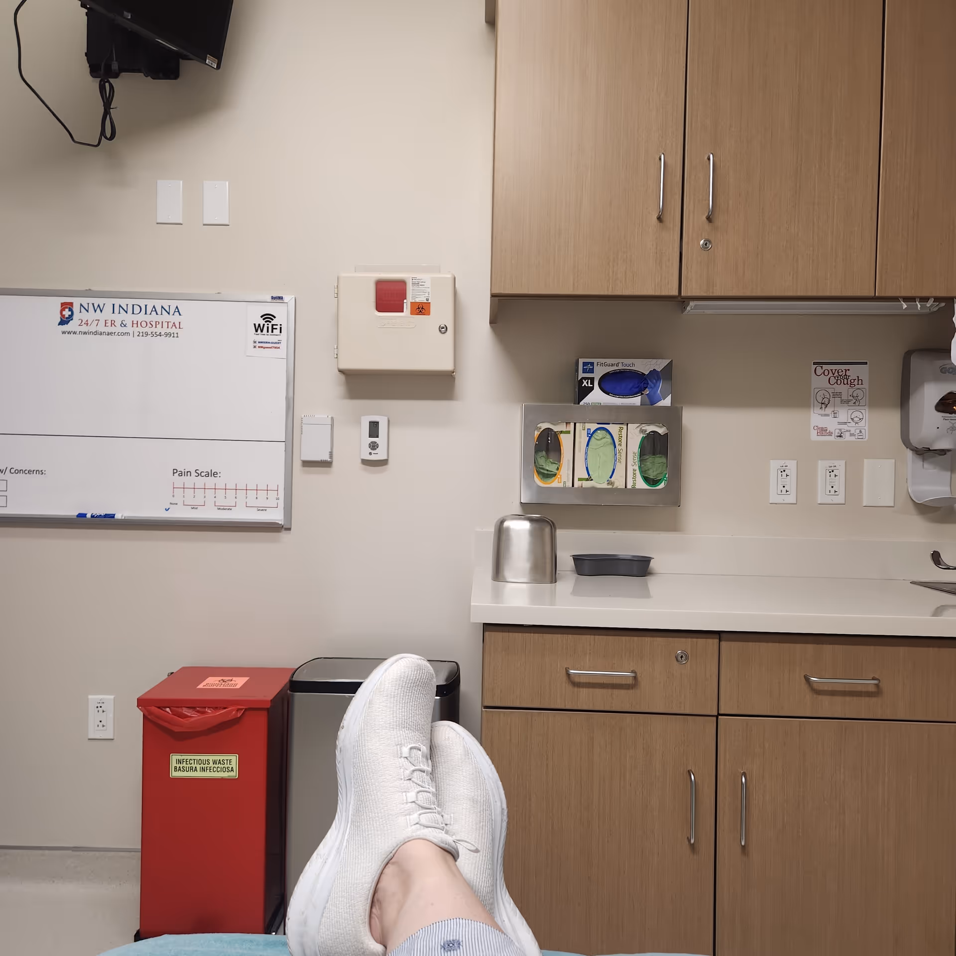 Feet in white sneakers rest on a chair in a hospital-like room with cabinets and whiteboard.