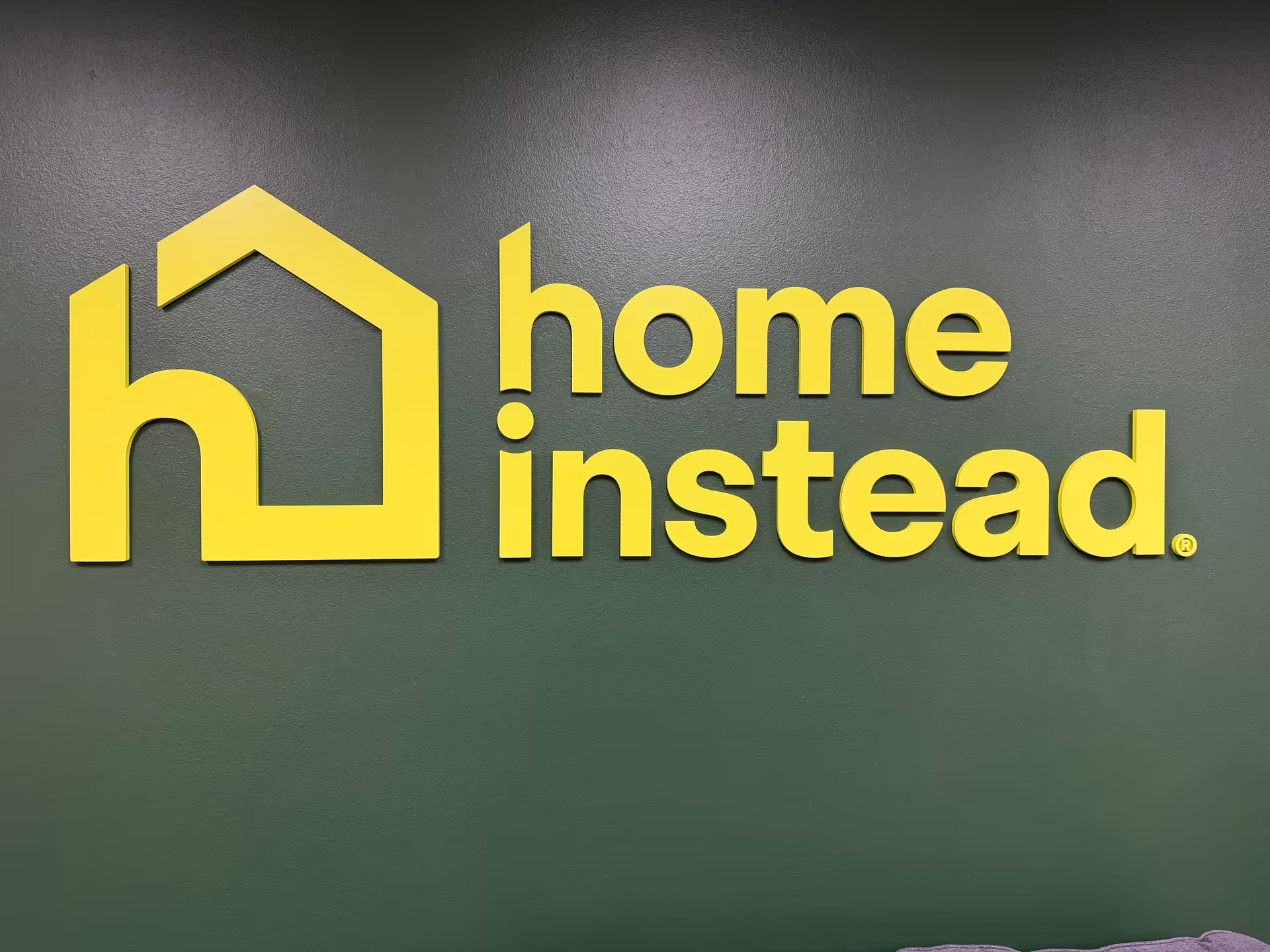 Bright yellow Home Instead logo mounted on a dark green interior wall.