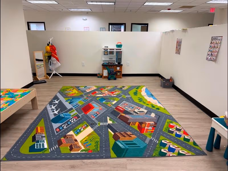 Colorful play mat with city streets occupies a therapy room, supplies and toys against white walls.