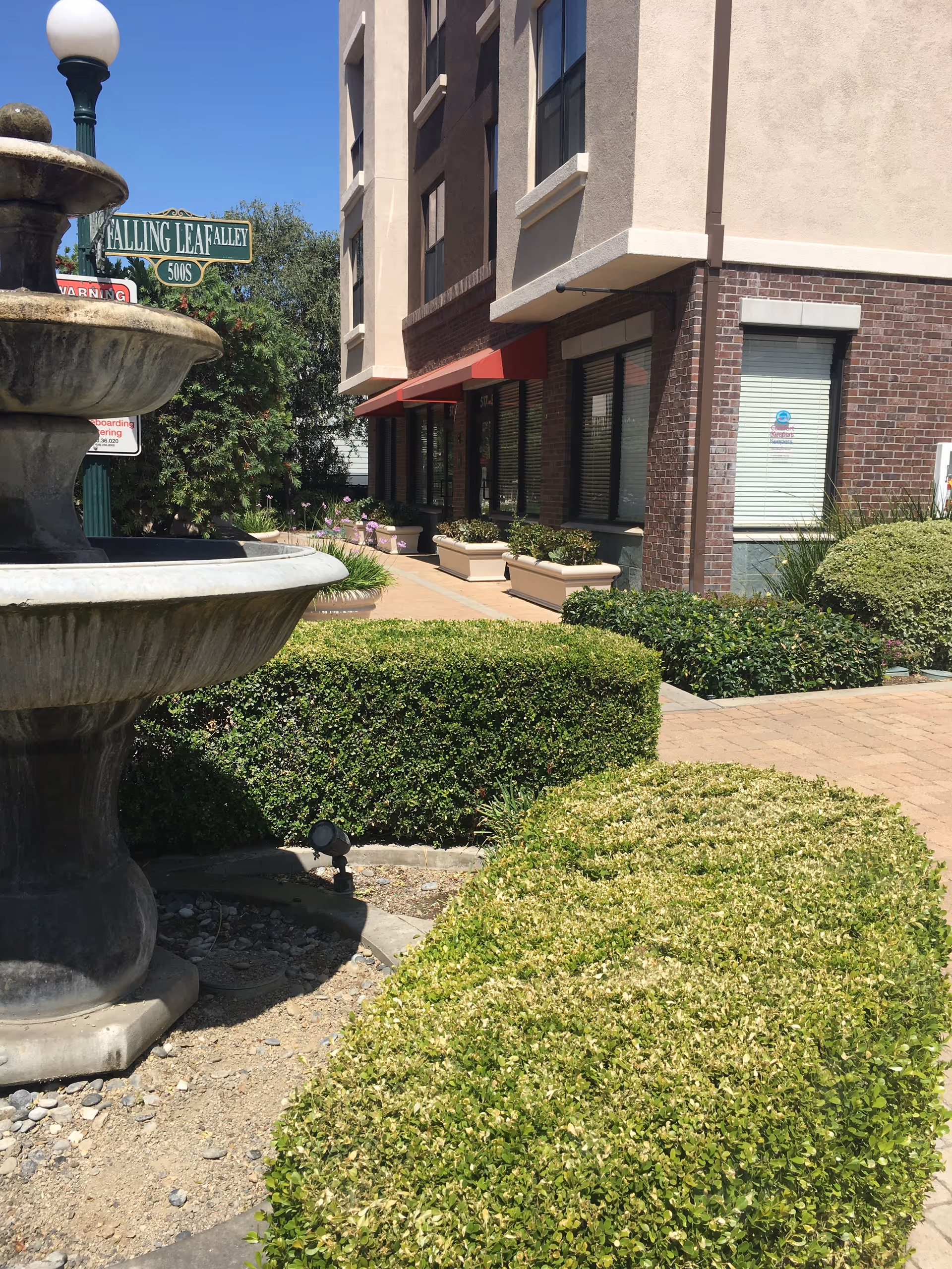Outdoor courtyard featuring a large fountain, brick building, planters, and trimmed hedges.