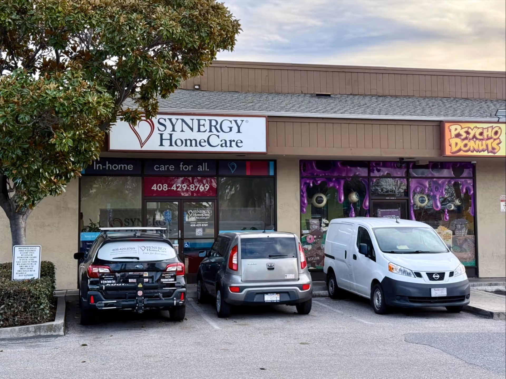 storefront of SYNERGY HomeCare in a strip mall with parked cars and a Psycho Donuts sign