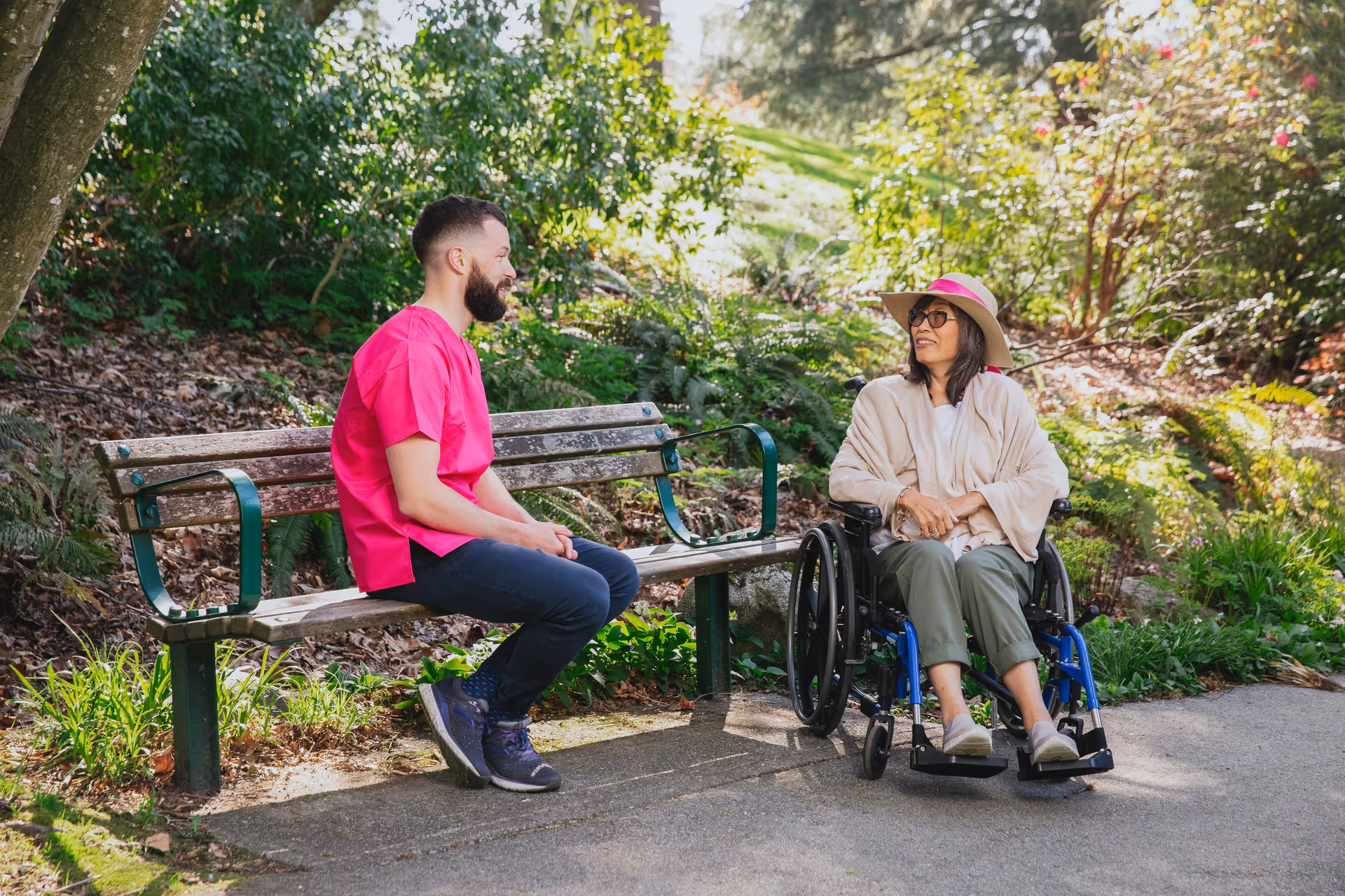 Caregiver in pink scrubs chats with a woman in a wheelchair on an outdoor bench.