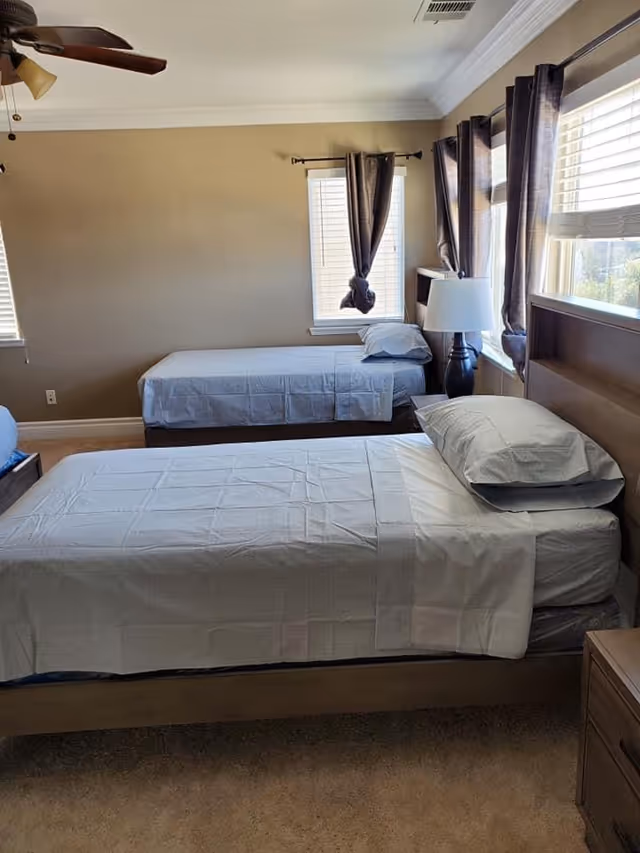 Two twin beds in a neutral senior living bedroom with lamps and windows.