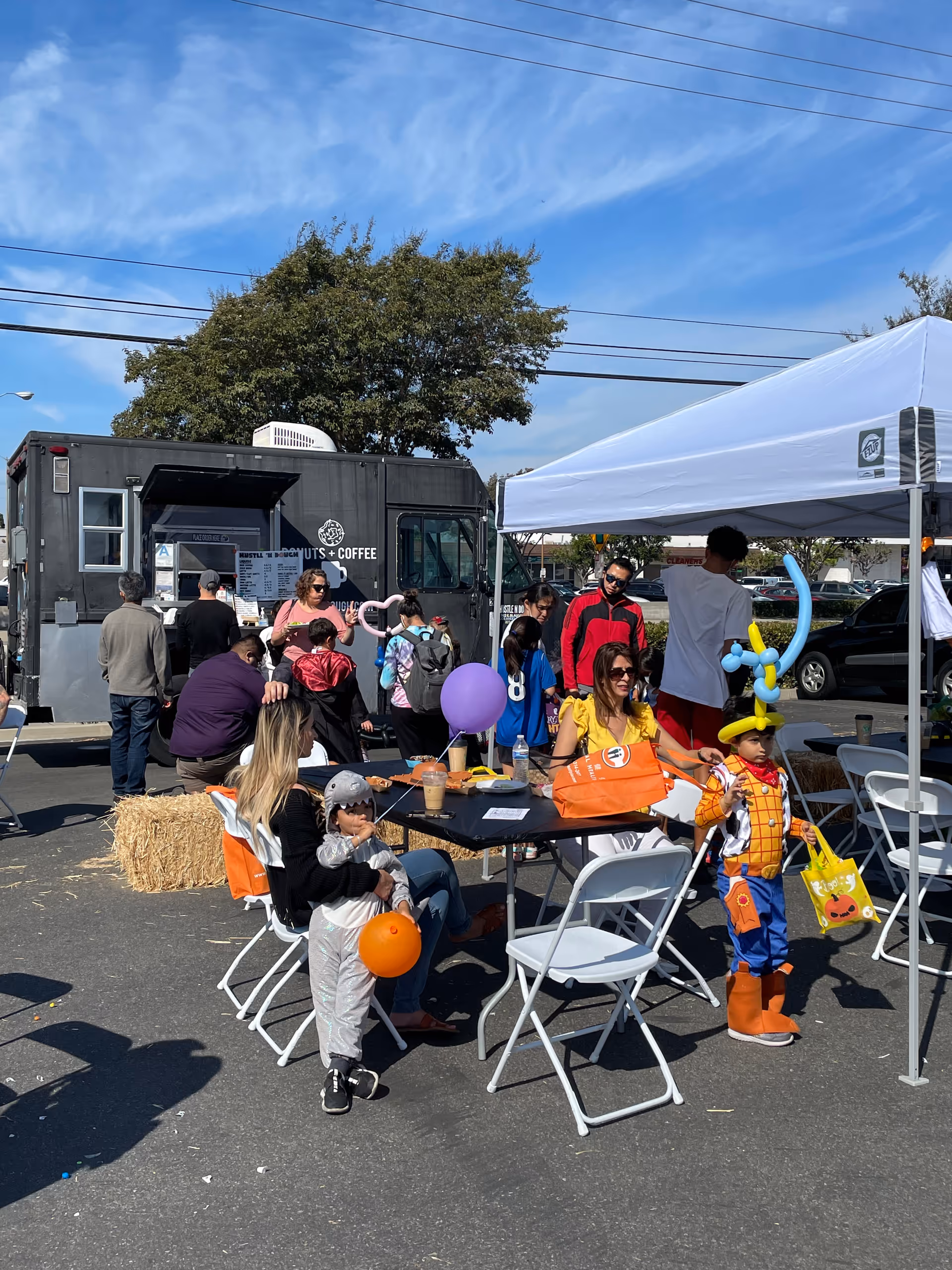 Outdoor community event at a senior facility with families, kids in costumes, balloons, and tables.