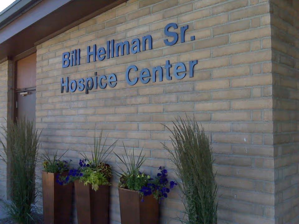 Exterior brick wall with Bill Hellman Sr. Hospice Center sign and tall potted plants.