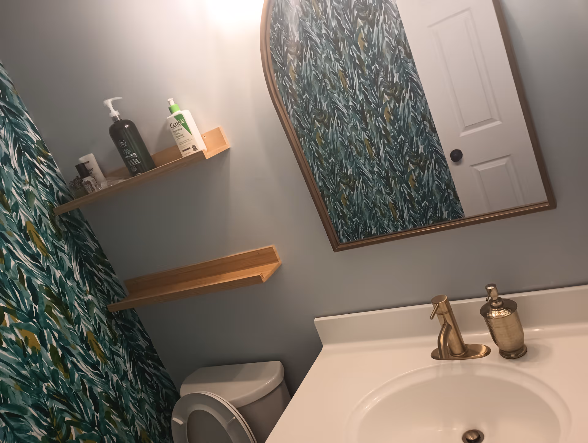 Bathroom with a tropical-pattern shower curtain, sink, mirror, toilet, and wall shelves.