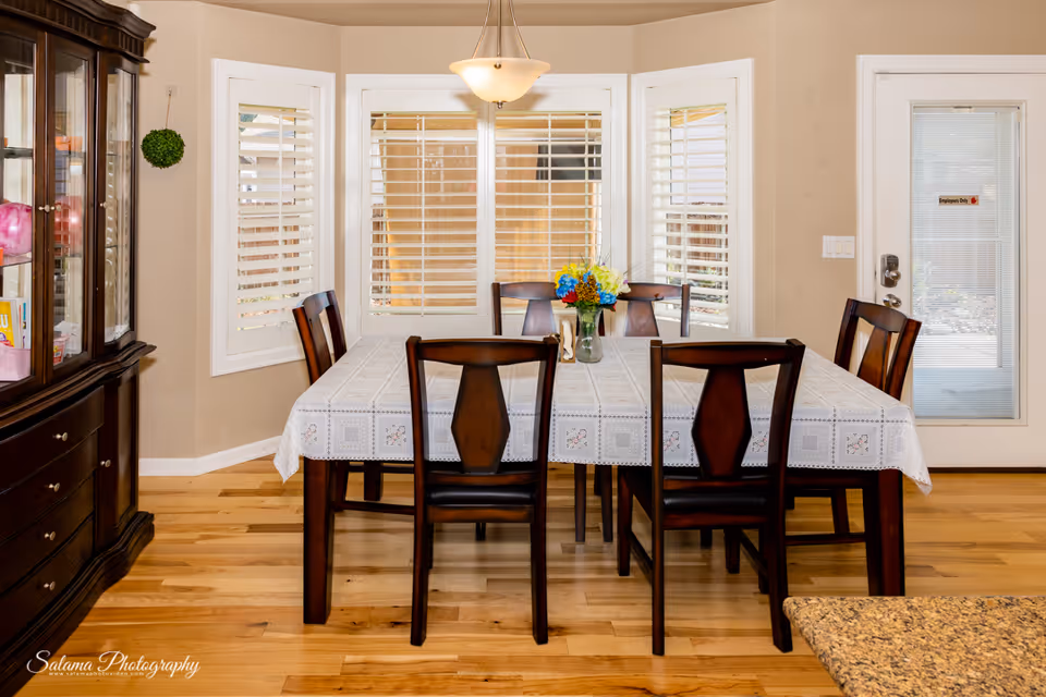 Bright dining room with table set for six, windows with blinds, and a china cabinet.