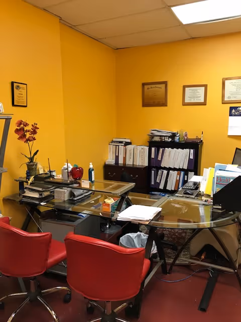 Yellow-walled office with glass desk, red chairs, and framed certificates.