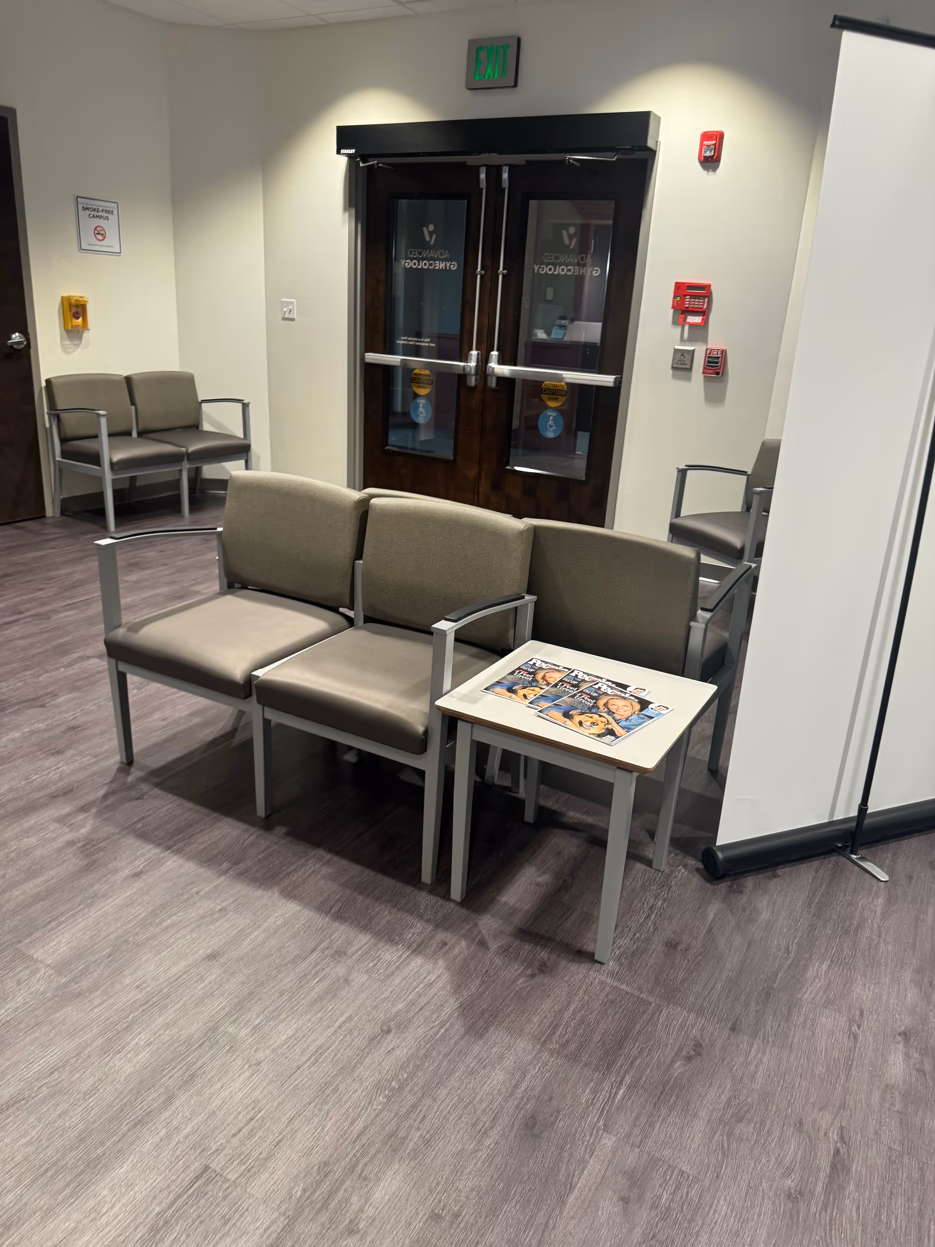 Waiting area with beige chairs, a magazine table, and double glass doors in a medical lobby.