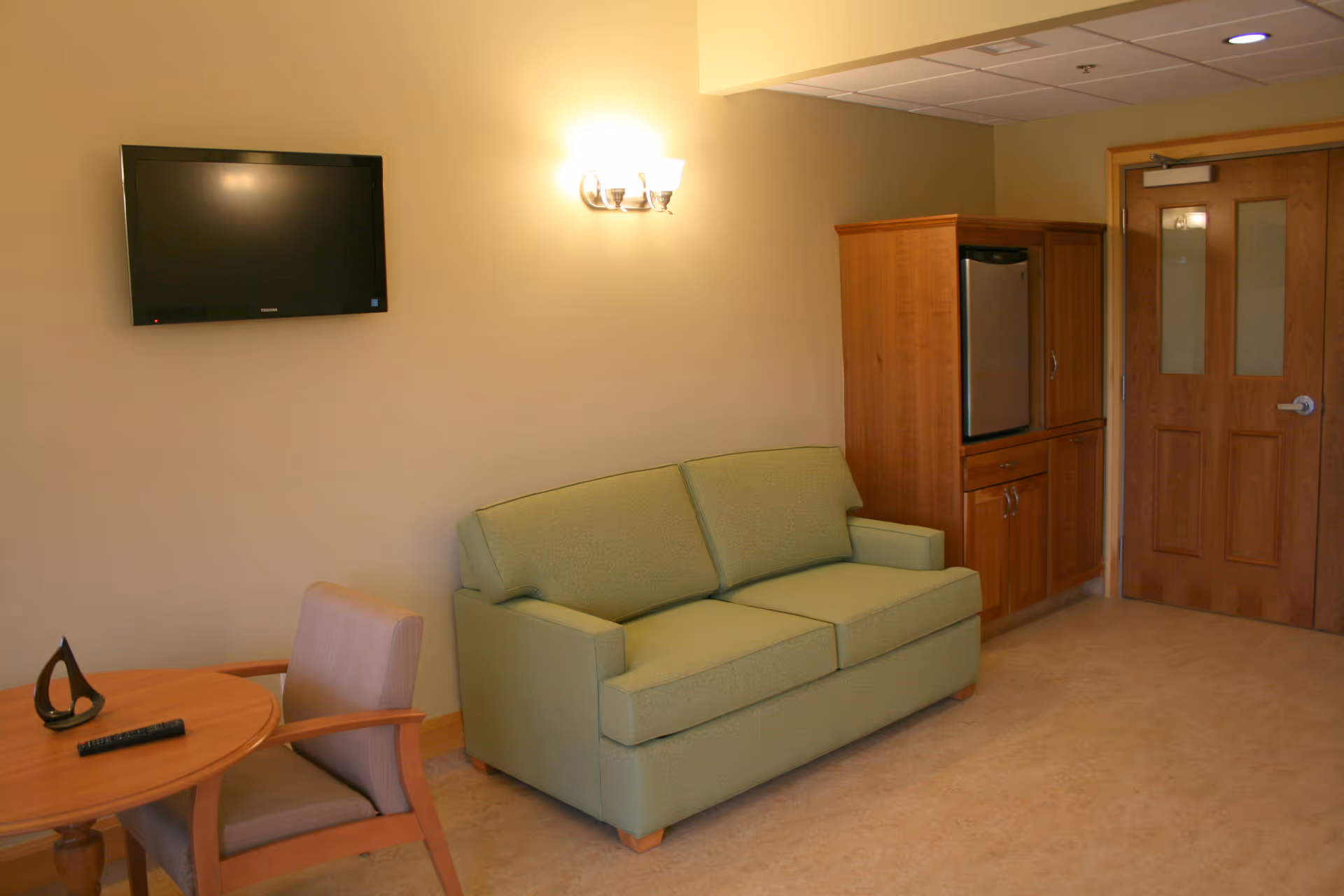 Lounge area with green sofa, wall TV, wooden cabinet, round table, and door.