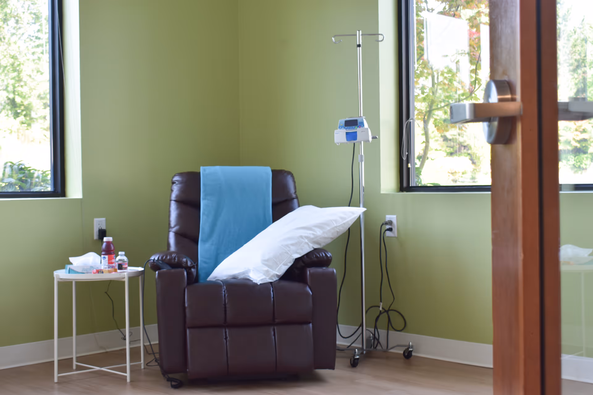 Green-walled infusion room with a brown leather recliner, IV pole, and side table with supplies.