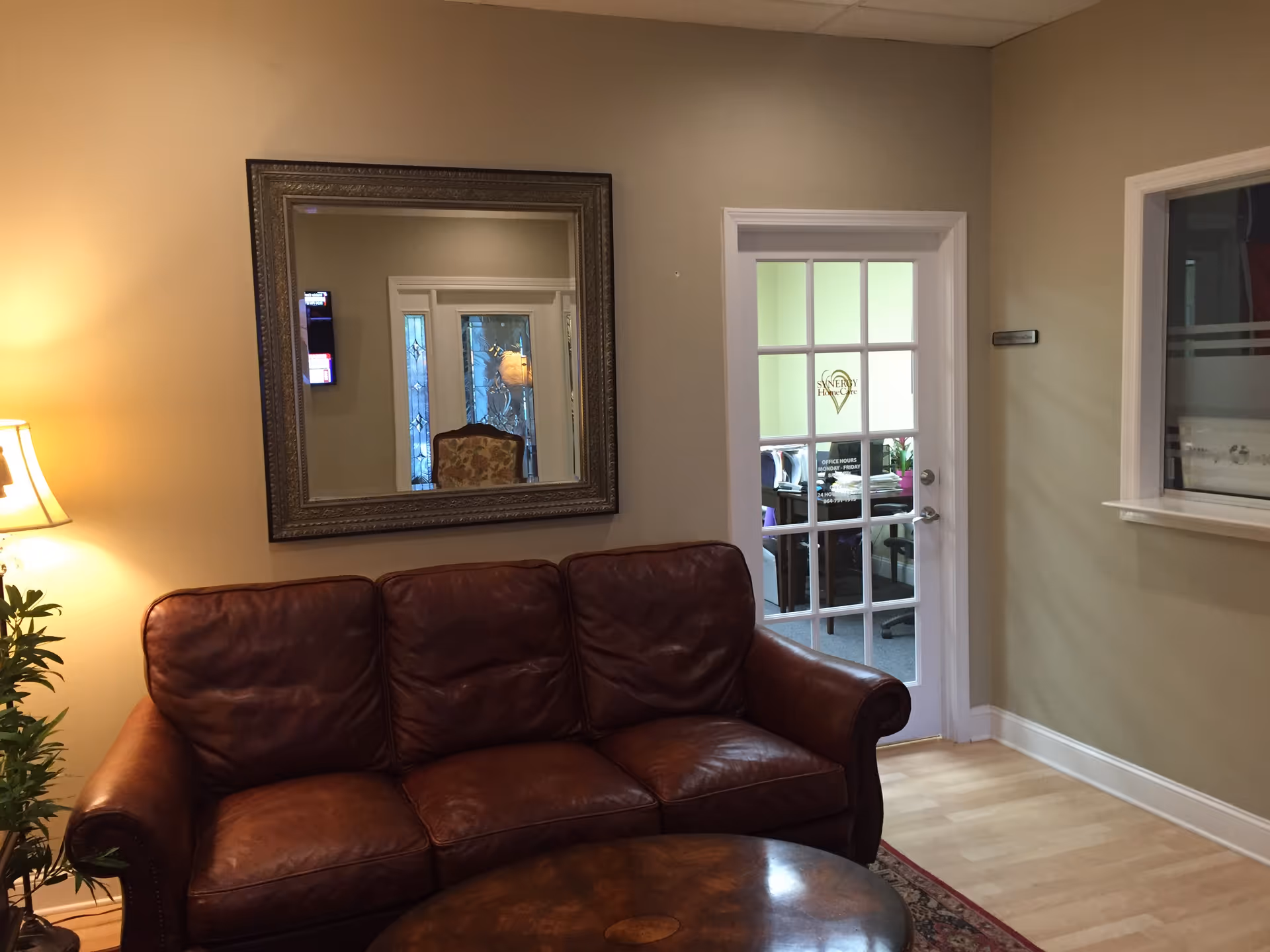 Cozy senior living lounge with leather sofa, lamp, large mirror, and glass-door entrance.