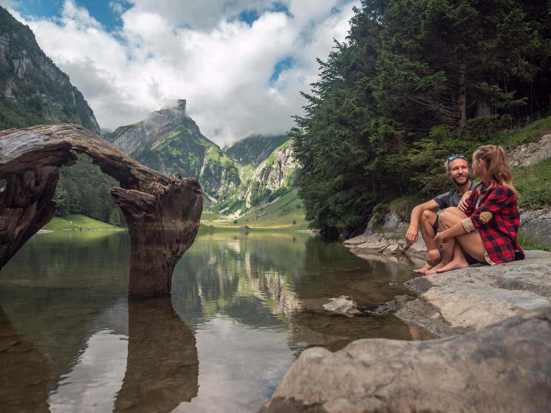 Two people sit barefoot on rocks beside a calm mountain lake.
