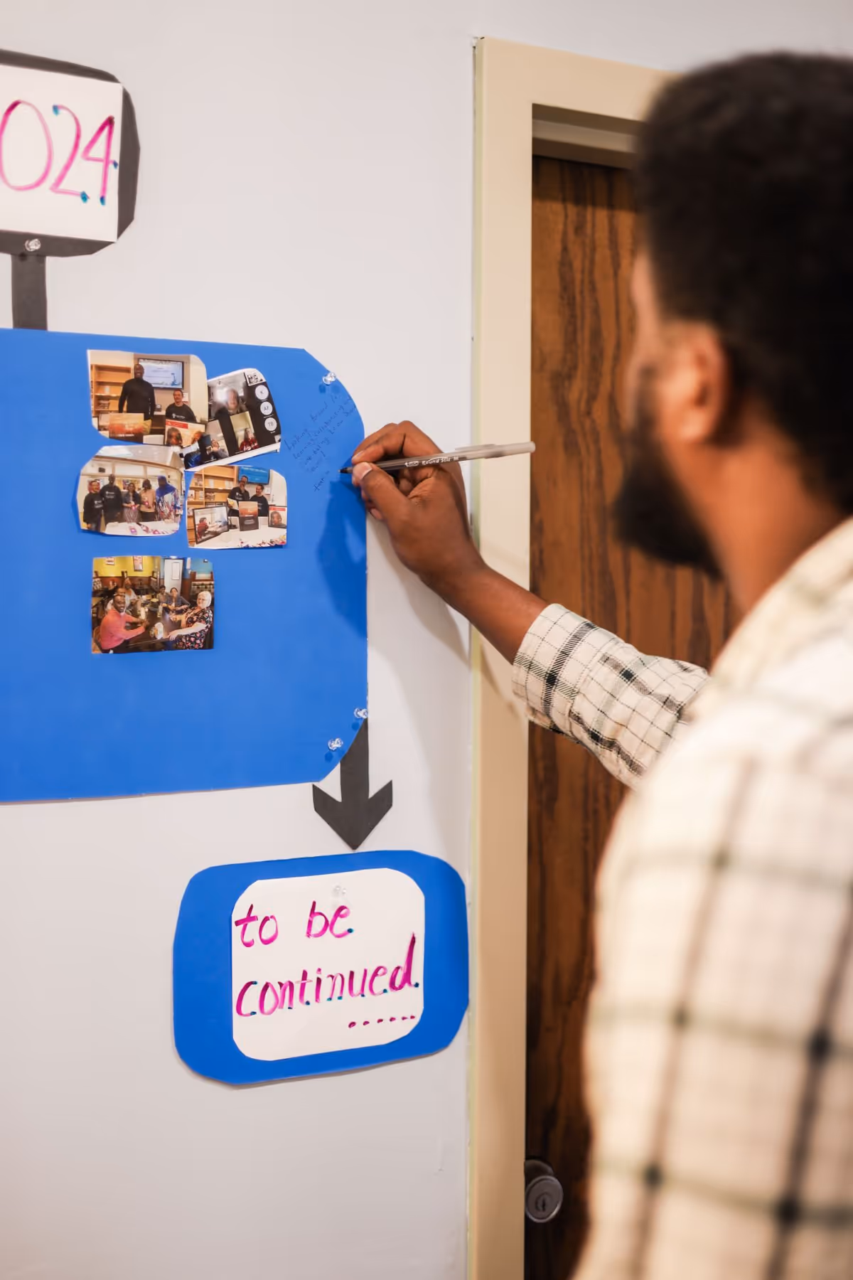 A man writes on a blue bulletin board in a hallway with photo collages.