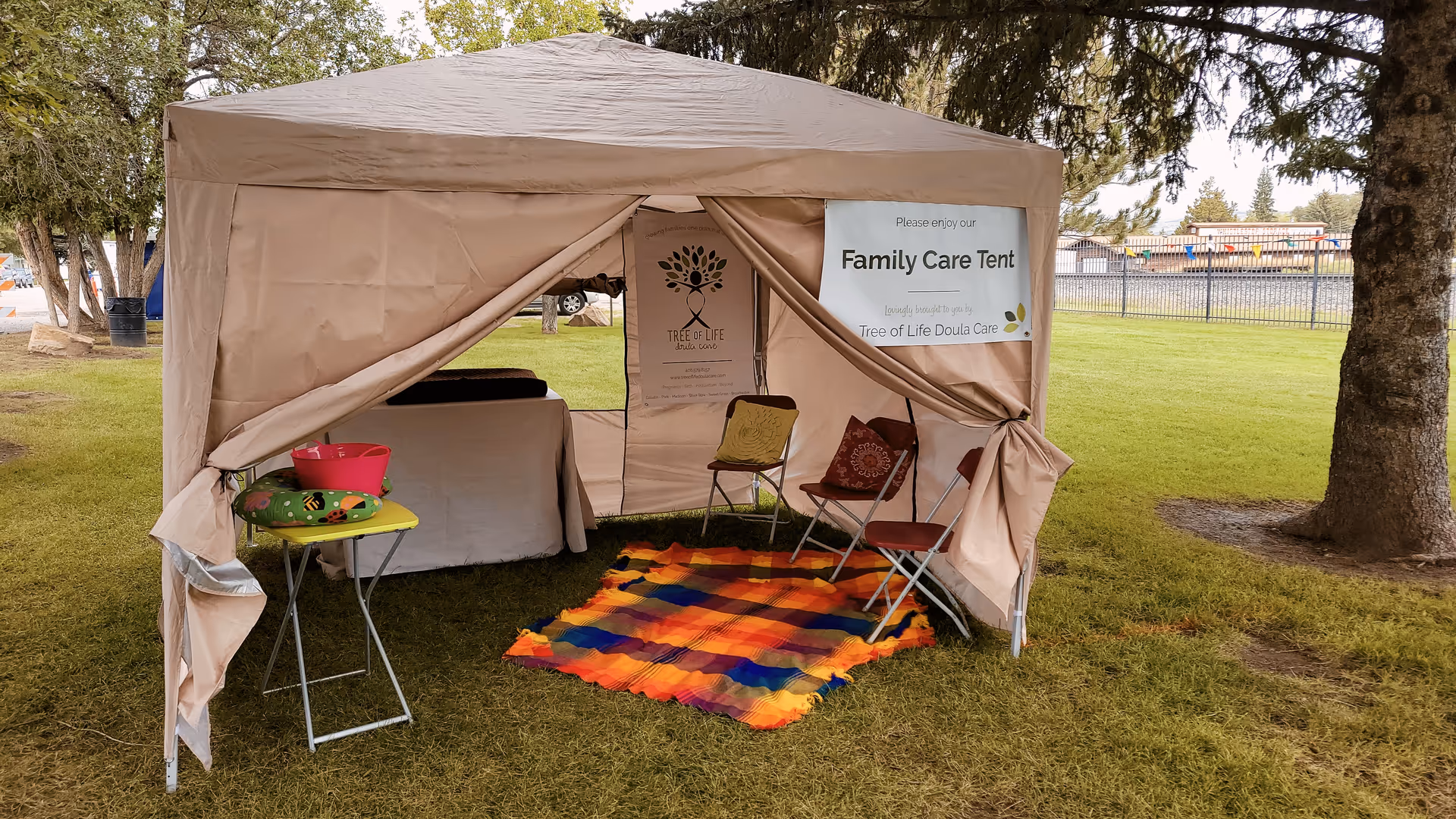 Open beige tent labeled Family Care Tent on grassy field with chairs and colorful rug.