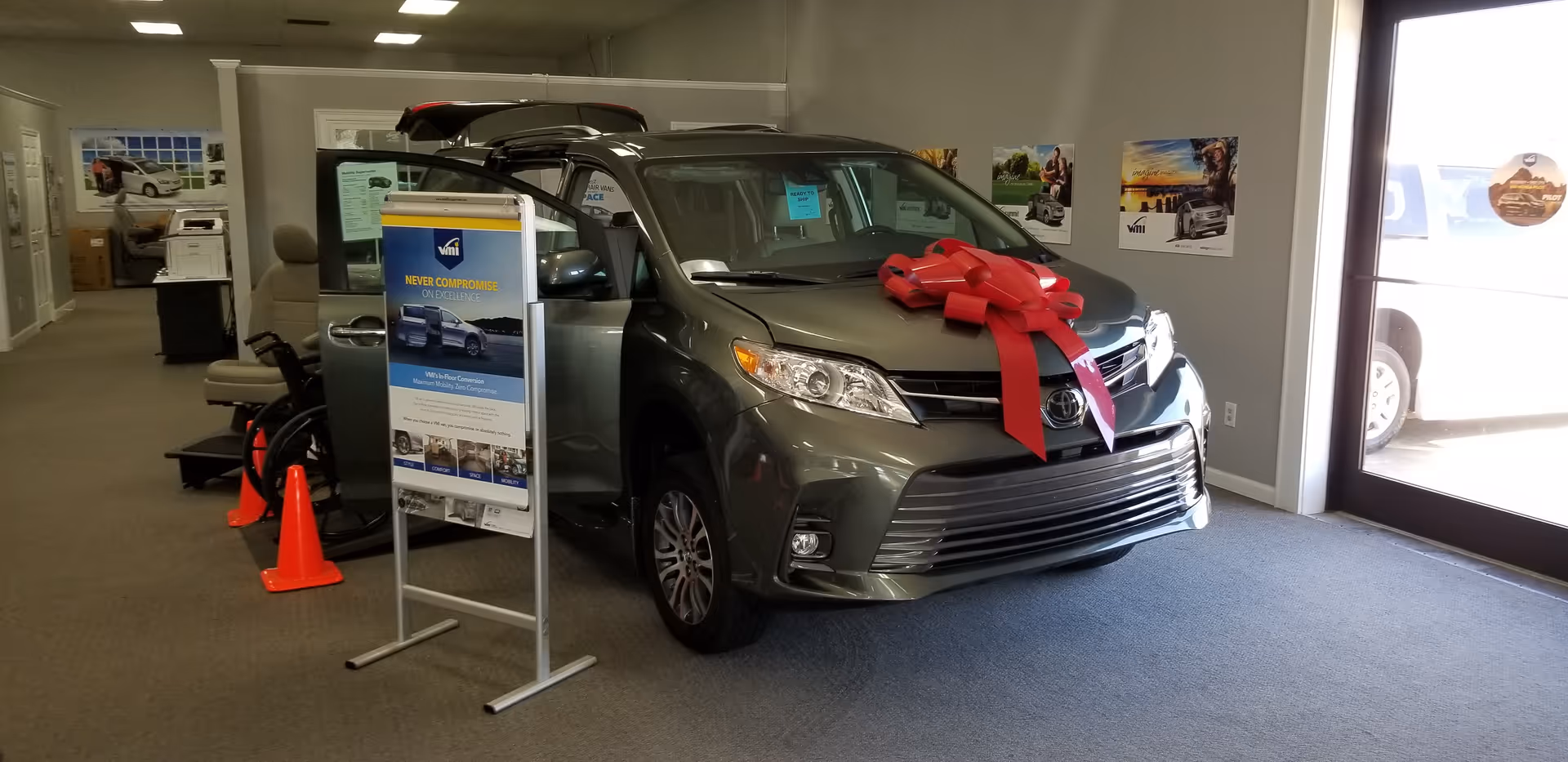 Inside a MobilityWorks showroom, a gray car with a red bow displayed beside posters.