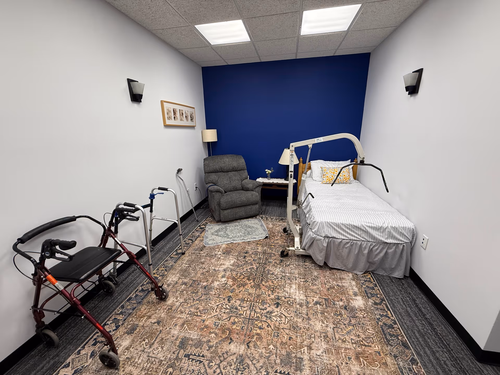 Bedroom-style medical room with adjustable bed, blue accent wall, lift frame, recliner, and walkers.