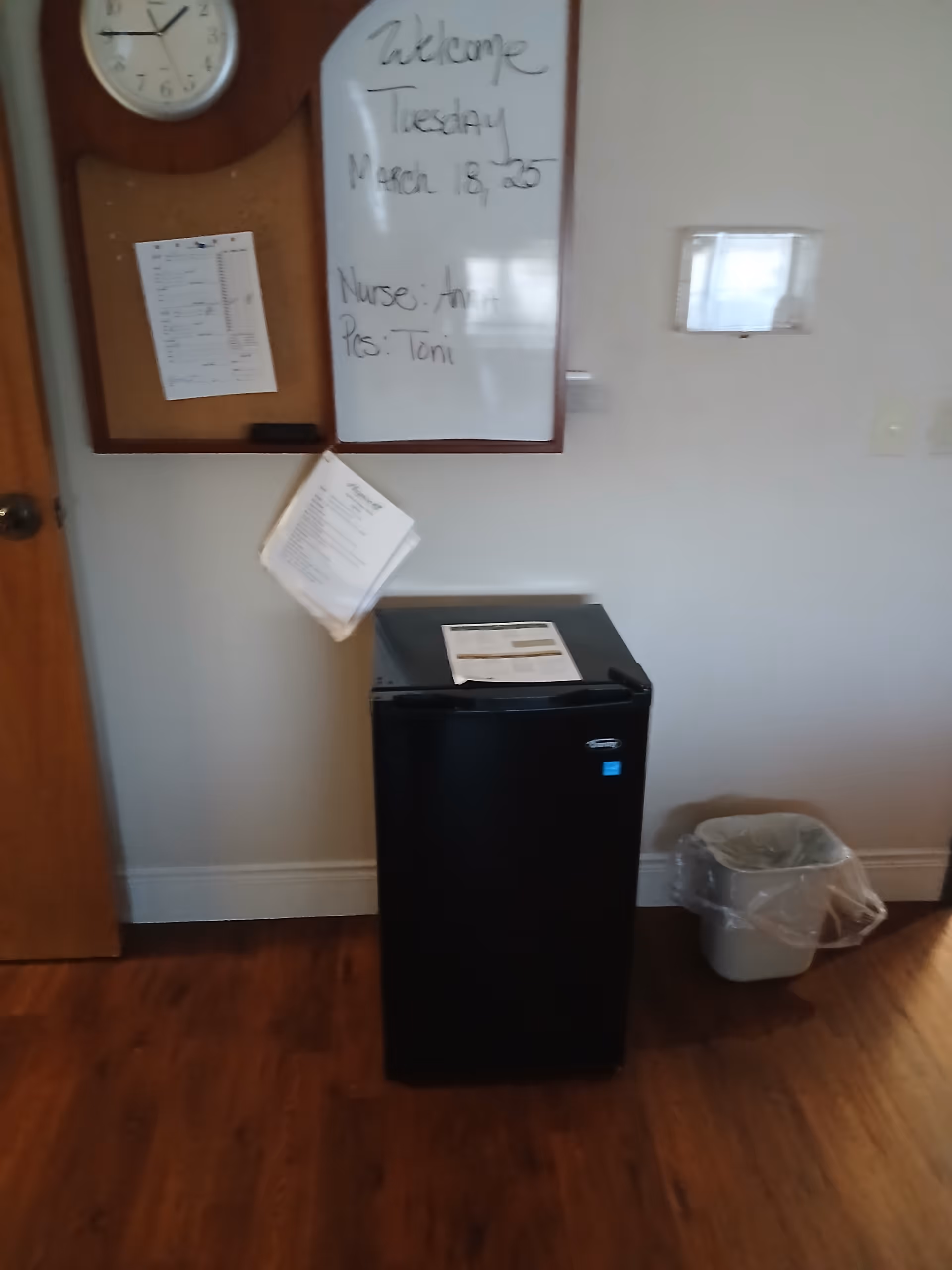 Interior common area featuring a whiteboard, clock, small fridge, papers, and a trash can.
